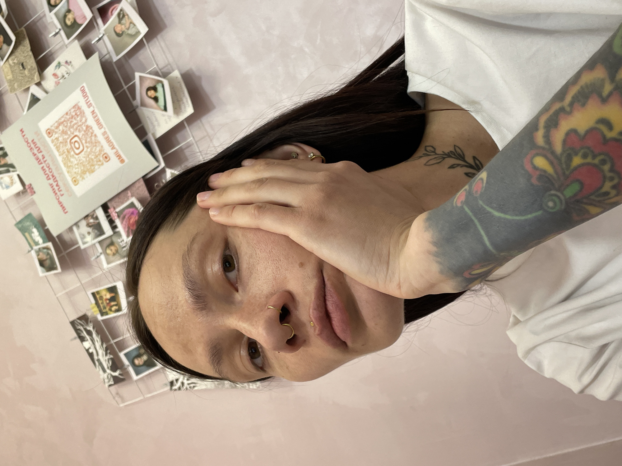 A person with dark hair, facial piercings, and a tattoo on their arm is looking at the camera. The person is wearing a white shirt. Behind them is a pink wall decorated with a wire grid holding various photographs and a card with a QR code and the text "@MILAGRES.GREEN.STUDIO" on it. The location is not evident from the image.