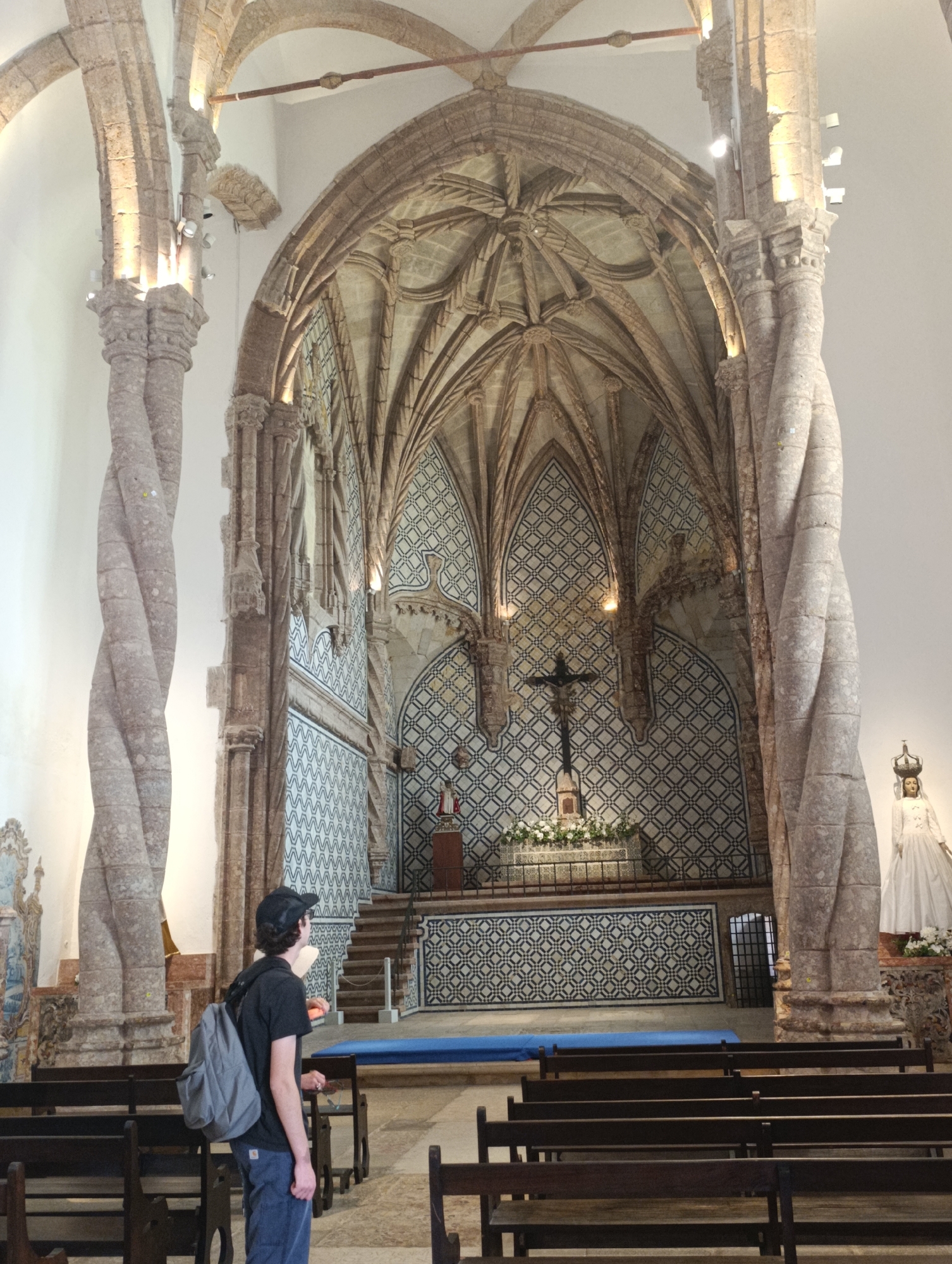 The image captures the interior of a church in Setúbal, Portugal, likely the Igreja de São Julião. A young person wearing a black t-shirt, blue jeans, and a black baseball cap stands facing away from the camera, with a gray backpack on their shoulders. They are positioned in the foreground, amidst rows of dark wooden pews.
The church's architecture is a prominent feature, with intricately carved stone pillars twisting upwards towards a vaulted ceiling. The walls are adorned with decorative tilework, featuring geometric patterns in blue and white. In the background, an altar area is visible, with a crucifix standing above a flower arrangement. To the right of the altar, a statue of a robed figure wearing a crown is present. The overall lighting suggests an indoor environment, with artificial lights illuminating the architectural details.