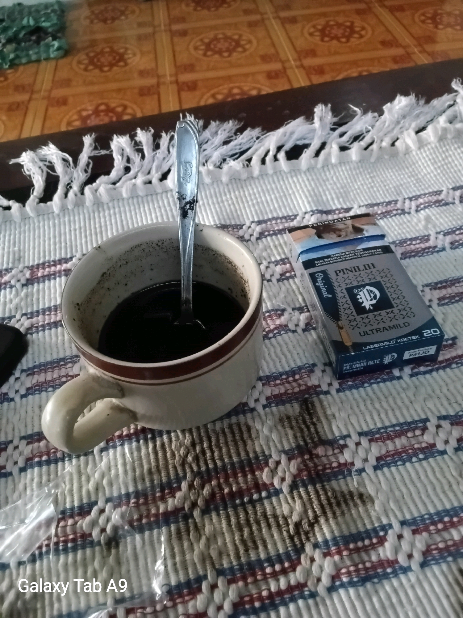 An indoor, high-angle shot shows a cup of dark coffee with a spoon in it, placed on a woven placemat. To the right of the coffee cup is a pack of "ULTRAMILD" cigarettes. The background includes a wooden table and a tiled floor with decorative patterns. The overall scene suggests a casual break or a moment of relaxation. The bottom left corner of the image displays a watermark that reads "Galaxy Tab A9". The setting appears to be a domestic or casual environment in Trenggalek, Indonesia.