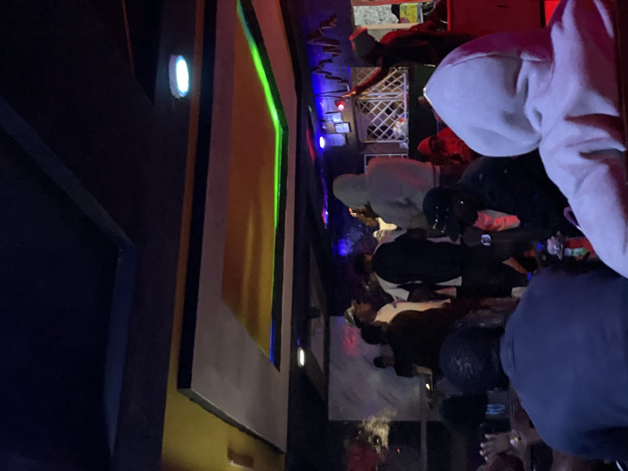 The image depicts an indoor scene, likely a social gathering or entertainment venue in Durumi, Nigeria. The environment is dimly lit, with a prominent ceiling featuring a multi-layered design of dark, yellow, and gray panels. Neon green and blue lights illuminate the edges of these panels, creating a vibrant, atmospheric glow. Several individuals are present, mostly standing or seated, with their backs partially or fully to the camera. In the foreground, a person wearing a light gray hooded sweatshirt is visible from behind. To their left, another individual in a dark jacket and cap is also seen from the back. Further into the scene, a group of people are gathered, some appearing to be talking or observing something. There are hints of what might be bar or counter area in the background, with what appear to be bottles and possibly a shisha pipe visible on a table. The overall impression is of a lively, possibly nocturnal social setting.