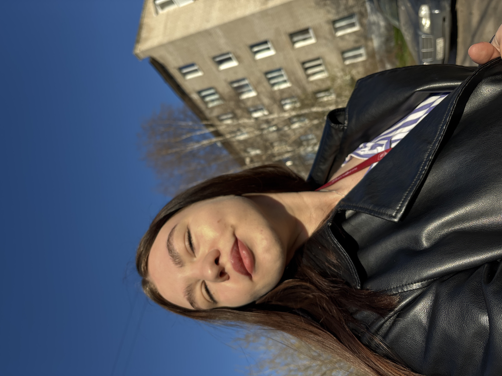 The image shows a young woman in a black leather jacket standing outdoors on a sunny day. She has long brown hair, fair skin, and her eyes are closed. Her lips are slightly parted, and her expression is relaxed. In the background, there is a tall, gray apartment building with many windows and a blue sky. A car is also partially visible in the background.
Based on the architectural style of the building and the overall environment, it is probable that this photo was taken in the city of Perm, Russia.