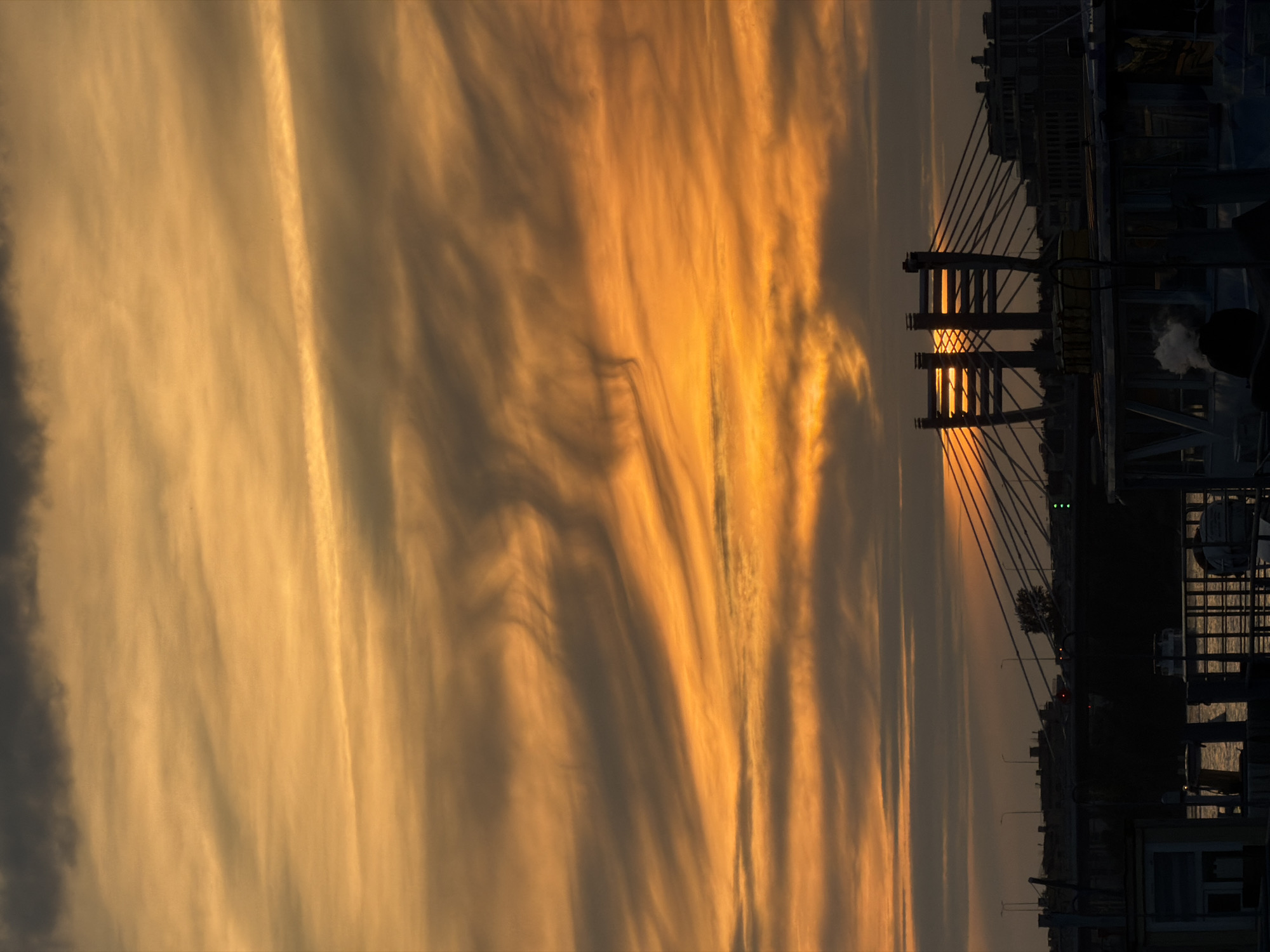 The image depicts a sunset over the Neva River in Saint Petersburg, Russia. The sky is filled with dramatic orange and gray clouds, illuminated by the setting sun. A cable-stayed bridge, likely the Bolshoy Obukhovsky Bridge, is silhouetted against the vibrant sky, with its distinctive pylons visible. In the foreground, the dark silhouettes of buildings and boats line the riverbank. A faint plume of smoke rises from one of the boats. The overall scene is one of a dramatic and atmospheric end to the day.