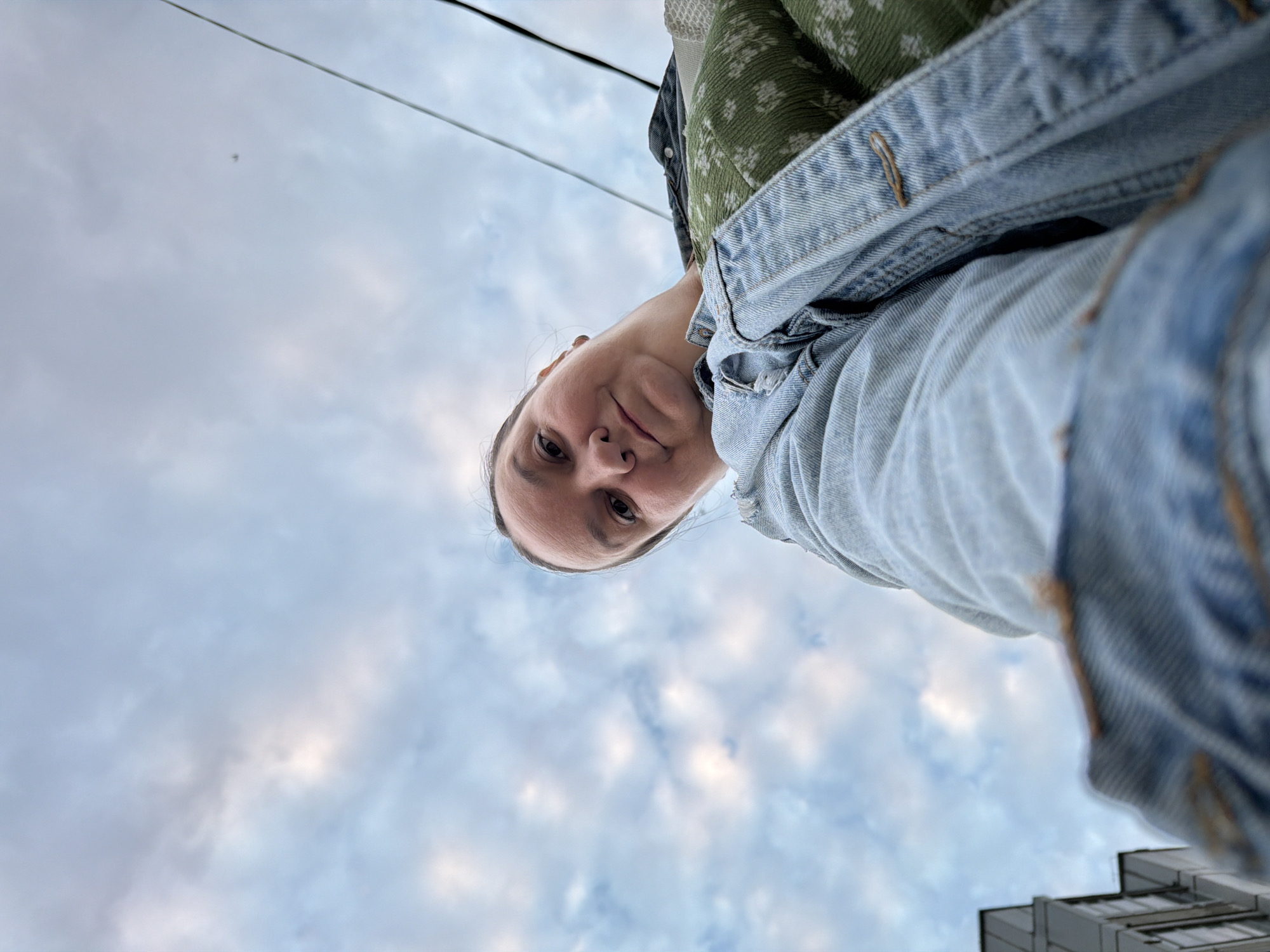 The image is a low-angle, wide-shot selfie taken outdoors. A woman with brown hair and fair skin is looking down at the camera. She is wearing a light blue denim jacket over a green patterned garment. The background consists of a cloudy sky and some wires. A building facade is visible on the left side of the frame. The perspective suggests the woman is standing and holding the camera at a low angle, possibly pointing upwards. The lighting is natural, indicating it is daytime. The overall scene is casual and appears to be taken in an urban or suburban environment, potentially in Chaikovsky, Russia, given the context.