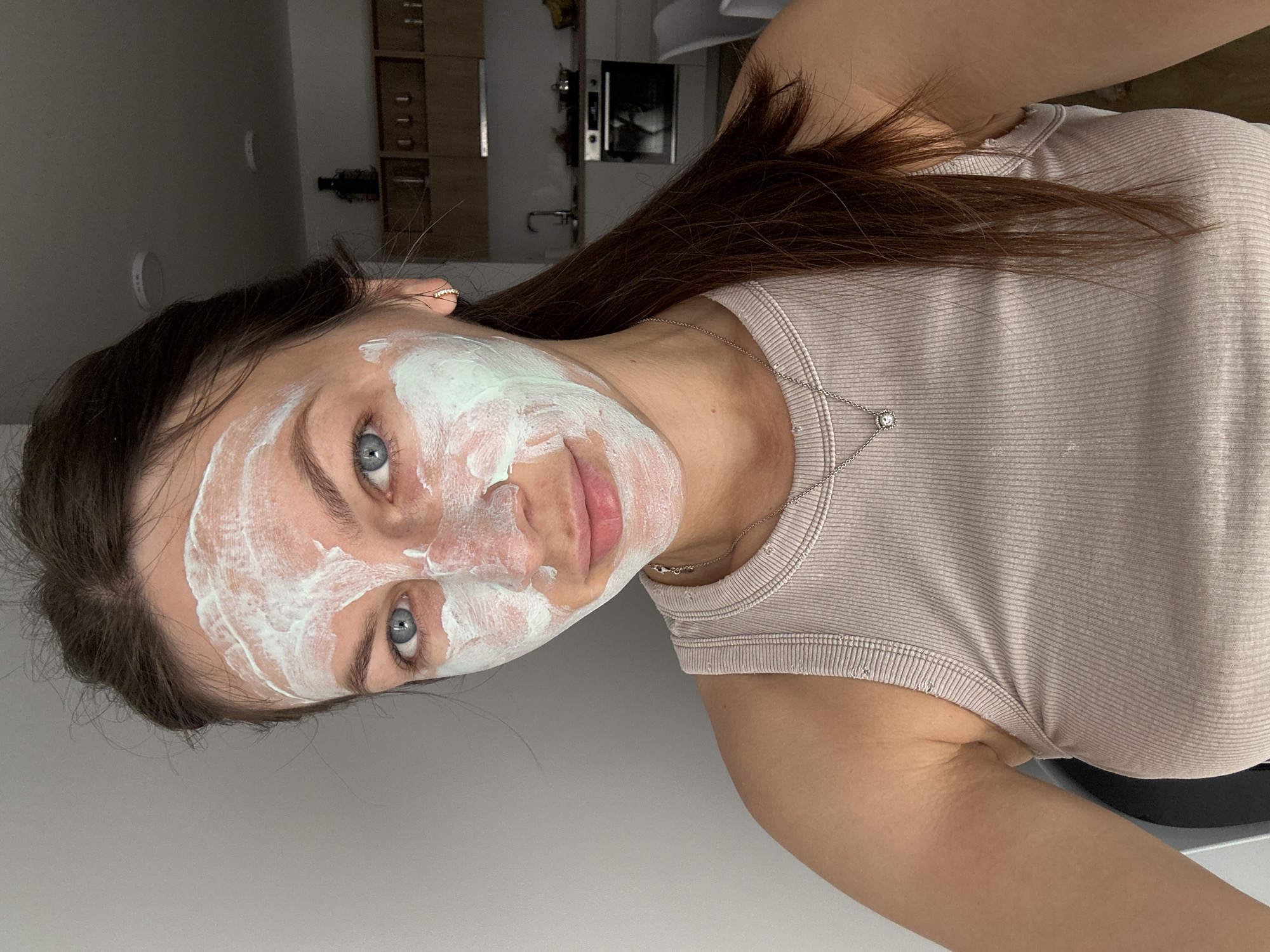 A woman with brown hair and blue eyes is wearing a green and white face mask. She is wearing a light-colored ribbed tank top and a silver necklace with a small pendant. The background shows a kitchen area with wooden cabinets, a sink, and a microwave. The location is implied to be Donji Pobori, Montenegro.