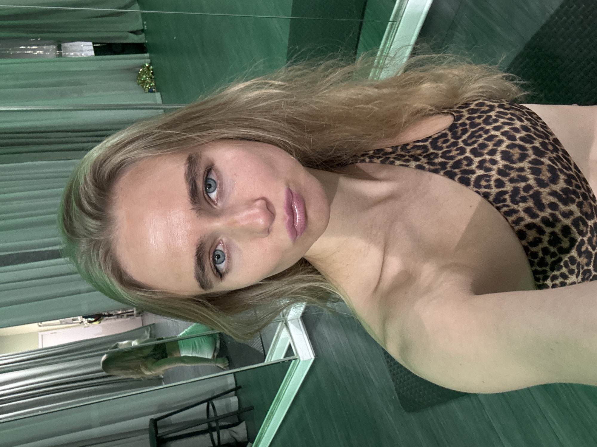 A fair-skinned female with blonde hair and blue eyes poses in what appears to be a gym or fitness studio in Unknown, Unknown. She is wearing a leopard-print sports bra. A mirror is behind her, reflecting a gym setting with workout equipment. The floor is green and textured. The overall lighting is bright.