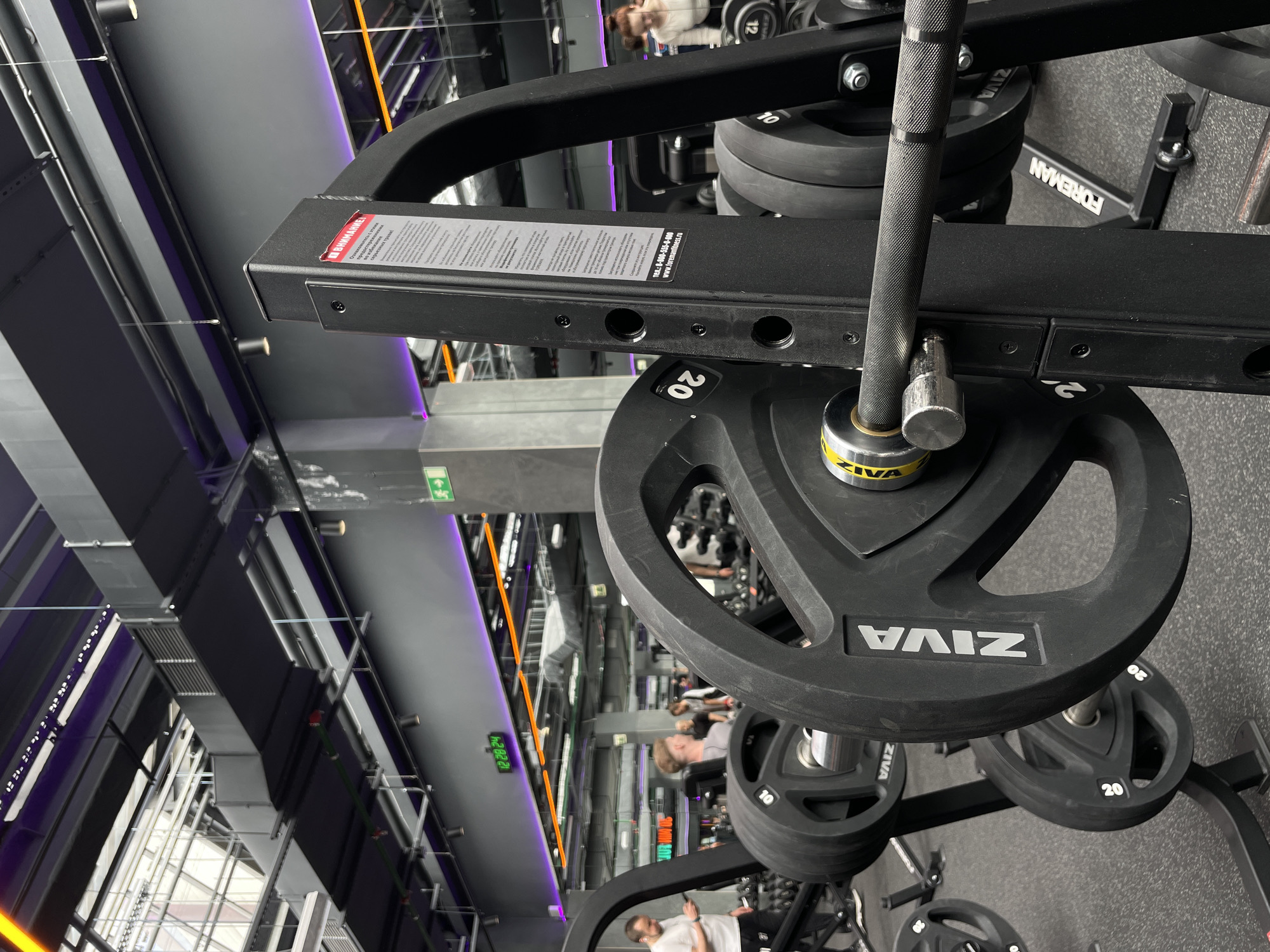 The image depicts the interior of a gym, specifically a weightlifting area. Weight plates, labeled "ZIVA" and marked with "20", "10", and "12" indicating their weight in kilograms, are loaded on a barbell supported by a squat rack. The rack, labeled "FOREMAN," has a warning sticker in Russian. The gym features a gray ceiling with exposed ductwork, linear lighting, and structural supports. People are visible in the background, engaged in fitness activities. The image likely originates from a gym located in the city of Kazan, Russia, given the Russian writing on the warning sticker.