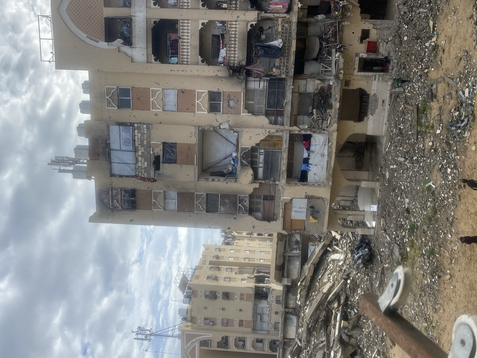 The image depicts an extensively damaged urban environment in Khan Yunis, Palestinian Territory, under an overcast sky. In the foreground, a rusted metal arm with two white pulleys extends from the lower left. Below it, a sandy-colored ground transitions into a vast expanse of rubble composed of concrete fragments, rebar, and debris. Three dark-clad individuals are walking across the sandy ground in the bottom left. Amidst the rubble, a dark, severely crushed vehicle is discernible.
Dominating the central and right portions of the frame is a multi-story, light-tan residential building exhibiting severe damage. Its facade is riddled with numerous impact marks, and many windows are shattered or entirely absent. Some window openings are covered with tarpaulins, a few of which display the UNICEF logo. Several balconies feature ornate balustrades but are partially destroyed, with laundry hanging from exposed interiors. Large sections of the building's walls are missing, revealing destroyed interior rooms. The ground level of this building is significantly damaged, with visible structural compromise and a dark-clad individual near an entry point.
To the left and rear, similar multi-story buildings, also appearing light-tan and heavily damaged, extend into the background. These buildings show numerous broken windows and signs of structural distress. A tall cellular antenna mast with multiple dishes is visible on the roof of a building in the far left background.