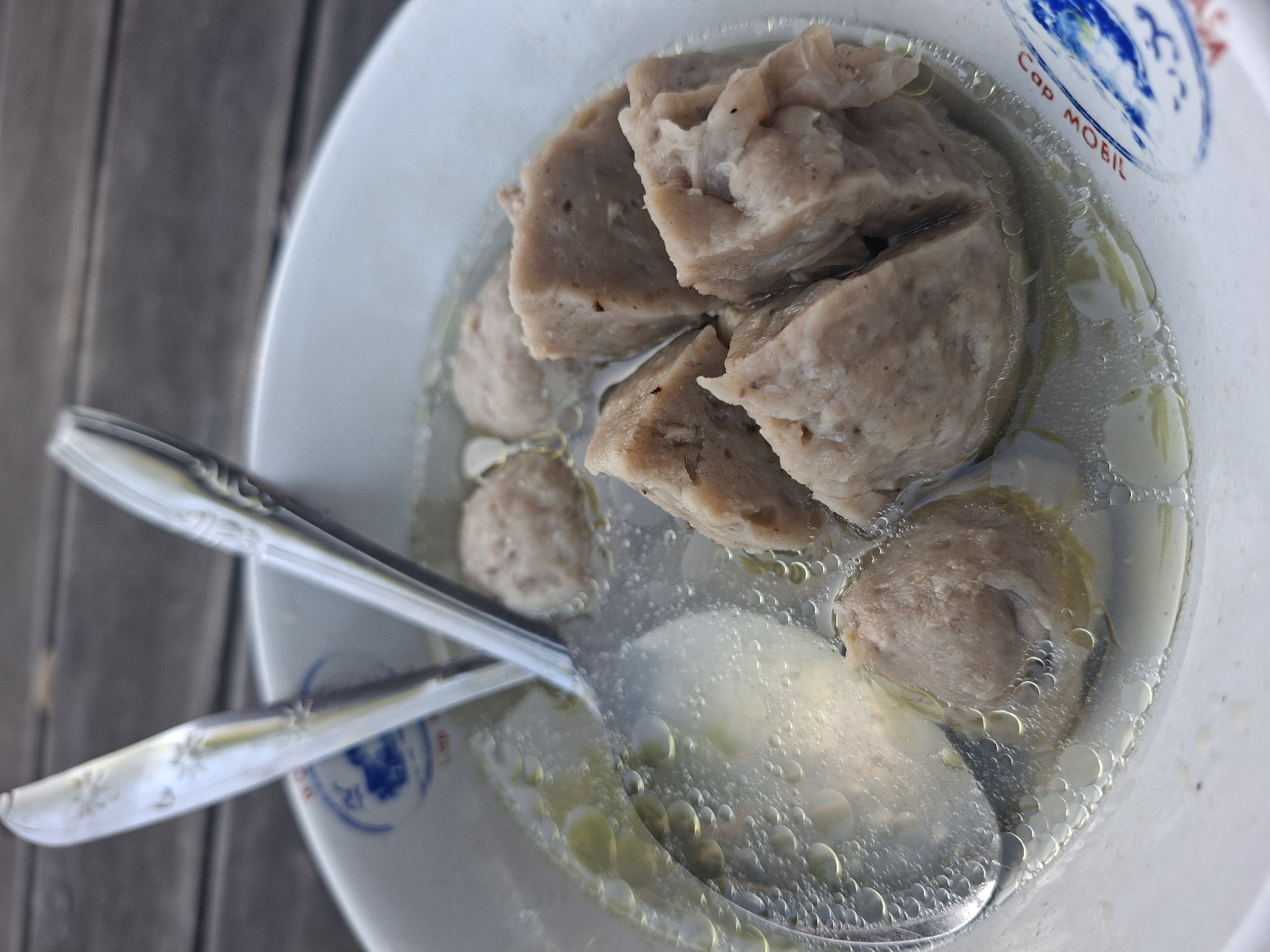 A close-up, top-down view shows a white bowl filled with broth and meatballs. Two silver spoons are resting in the bowl, with one spoon partially submerged in the broth. The broth is clear and slightly oily, with small bubbles on the surface. Several meatballs, some round and others cut into cubes, are visible in the broth. The bowl has blue and white insignia on the rim. The background appears to be a wooden surface, possibly a table. The image is shot outdoors, likely in a restaurant or food stall setting in Gianyar, Indonesia, indicated by the text "Cap Mobil" on the bowl rim.
