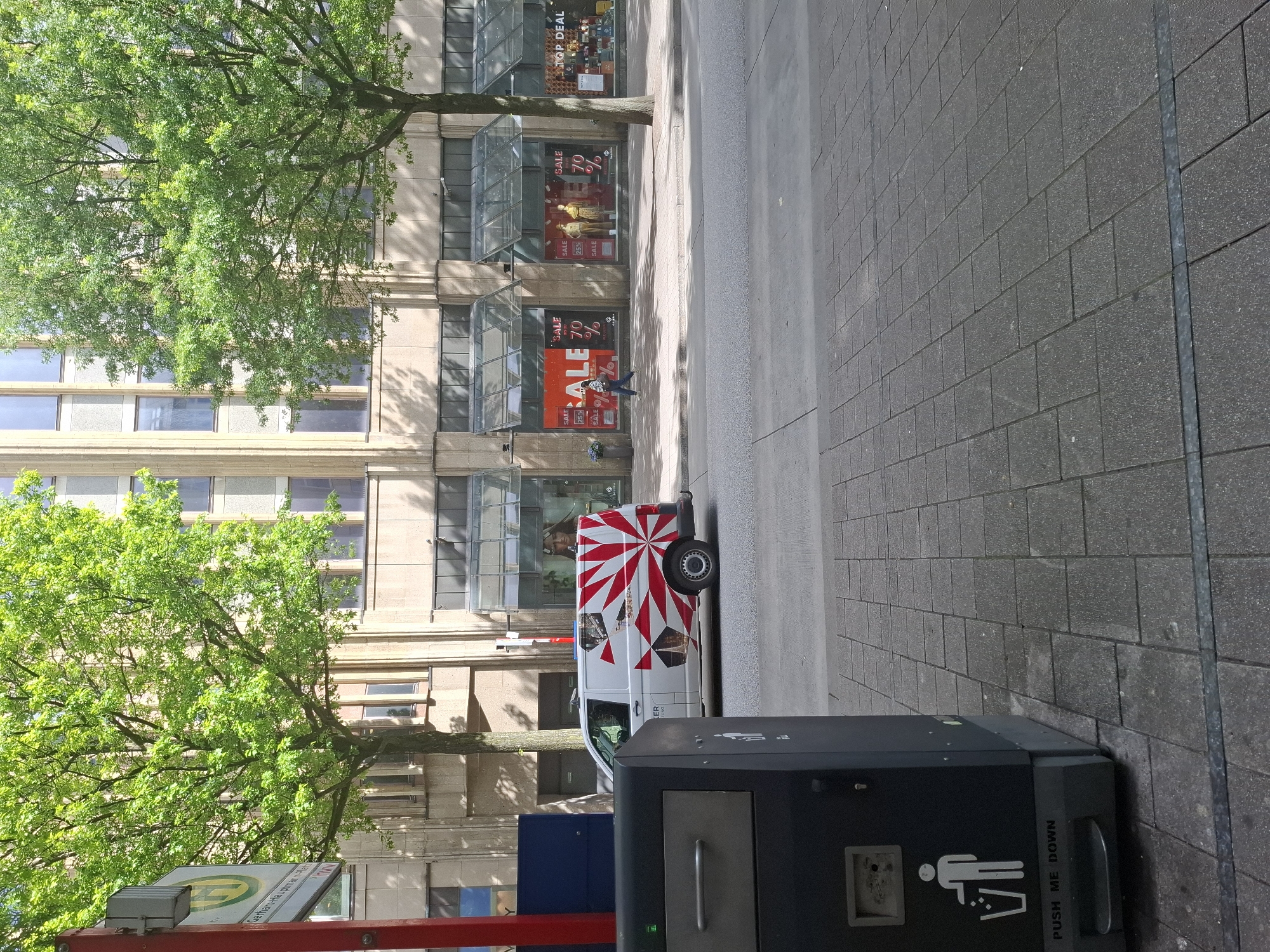 The image shows a street scene in Hamburg, Germany, likely in an urban commercial area. In the foreground, a black trash receptacle with a metal handle and a "Push Me Down" label is visible on a grey tiled sidewalk. To the right of the trash can, a white van with a distinctive red and white sunburst graphic and images of landmarks is parked on the street. Behind the van, a row of buildings with large glass storefronts displaying "SALE" signs with percentages like 25% and 70% are visible. A person in dark clothing is walking on the sidewalk in front of these storefronts. Tall trees with lush green foliage line the sidewalk, casting shadows on the pavement. The overall impression is a daytime scene in a well-maintained urban environment.