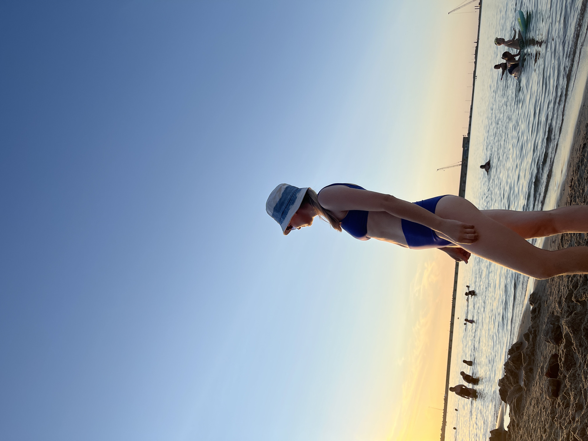 The image depicts a beach scene in the city of Pionersky, Russia, during what appears to be sunset or early evening. The sky is a clear gradient of pale blue to a faint yellow-orange near the horizon. In the foreground, a sandy beach is visible with some scattered rocks.
Dominating the right side of the frame is a young woman wearing a blue bikini and a denim bucket hat. She is facing right, in profile, looking out towards the water. Her pose suggests she is standing on the beach.
In the middle ground, the sea is calm with gentle ripples. Several people are in the water, some standing and others swimming or playing. To the left, there's a group of people wading near the shore. Further out, there are more individuals scattered across the water, and a few are on what appear to be paddleboards or inflatable devices.
In the background, a long pier or breakwater stretches across the water. Two construction cranes are visible on the horizon, suggesting ongoing or recent development along the coastline. The overall atmosphere is serene and recreational, typical of a beach at the end of the day.