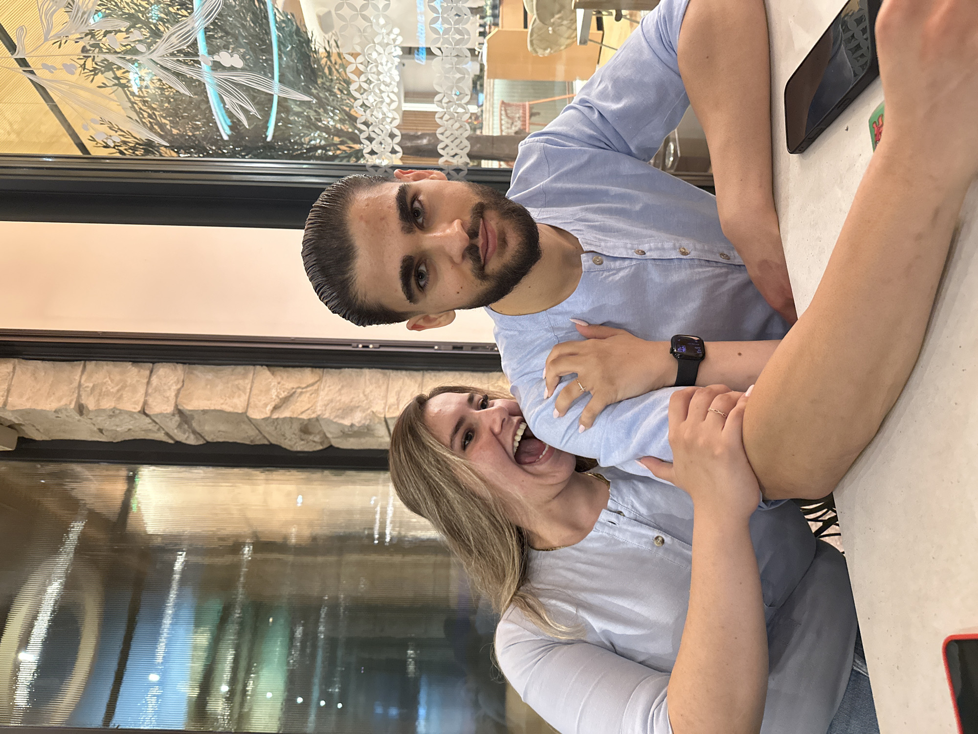 In Nicosia, Cyprus, a medium shot captures a young Caucasian couple sitting at a table. The woman, on the left, has blonde hair and is leaning towards the man with her mouth open in a laugh, her right arm around his shoulder and left hand resting on his arm. She wears a light blue button-down shirt and a delicate ring on her left hand. The man, on the right, has dark hair slicked back, a beard, and is looking directly at the camera with a neutral expression. He wears a similar light blue button-down shirt and a smartwatch on his left wrist. A smartphone lies on the table in front of him. The background features a large window with vertical ridges and a stone-clad wall. The interior of the establishment, visible through the window, shows decorative elements and other patrons. The scene suggests a casual dining or social setting.