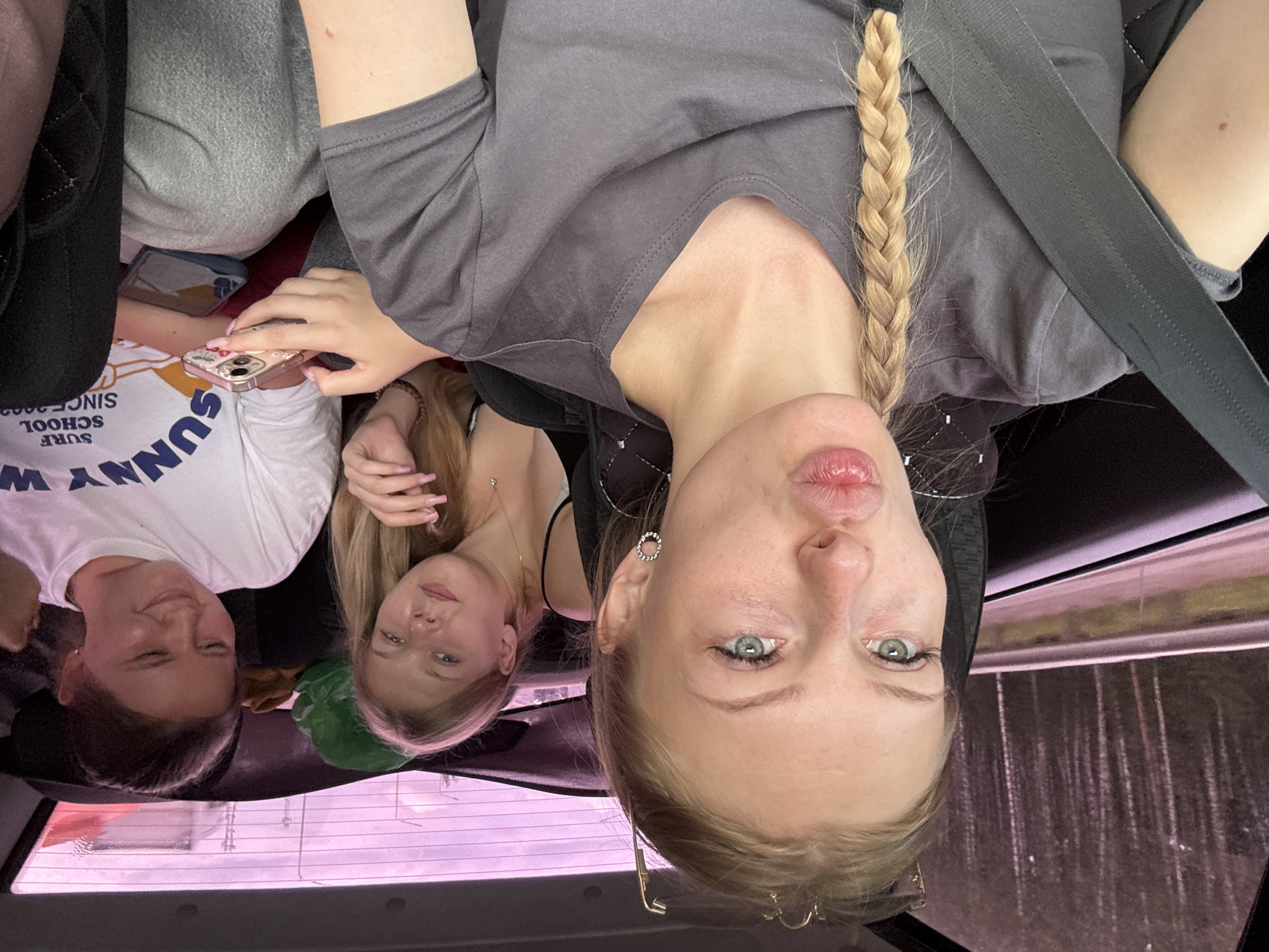 Three young women are inside a car, likely traveling on a road. The woman in the foreground is taking a selfie, puckering her lips and looking towards the camera. Her right arm is resting on her lap, and a seatbelt is visible across her chest. Her hair is blonde and styled in a braid that falls over her shoulder. She is wearing a dark grey t-shirt.
To her left and slightly behind her, another young woman with blonde hair is looking towards the camera with a relaxed expression. She is wearing a black top.
To the right and further back, a third young woman with dark hair is looking down at her phone, which she is holding in her hands. She is wearing a white t-shirt with "SUNNY SCHOOL" printed on it. Two mobile phones are visible in the image.
The car's interior is visible, including the car seats and the rear window, which is tinted pink and shows trees and sky outside, suggesting a forested or natural environment. The overall atmosphere appears casual and informal, consistent with a road trip or outing. There is no specific indication of Beloborodovo, Russia in the image, but the presence of a road and trees could align with a rural or suburban setting in Russia.