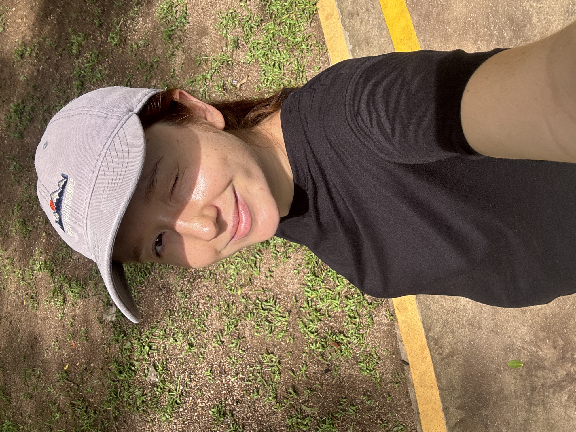 A light-skinned person with reddish-brown hair is outdoors. They are wearing a dark-colored t-shirt and a light-colored baseball cap with a small mountain-themed logo. The person is smiling and winking at the camera. The background consists of grass and a concrete surface with a yellow painted line. The image appears to be a selfie. The location is consistent with outdoor settings in Choeng Thale, Thailand.