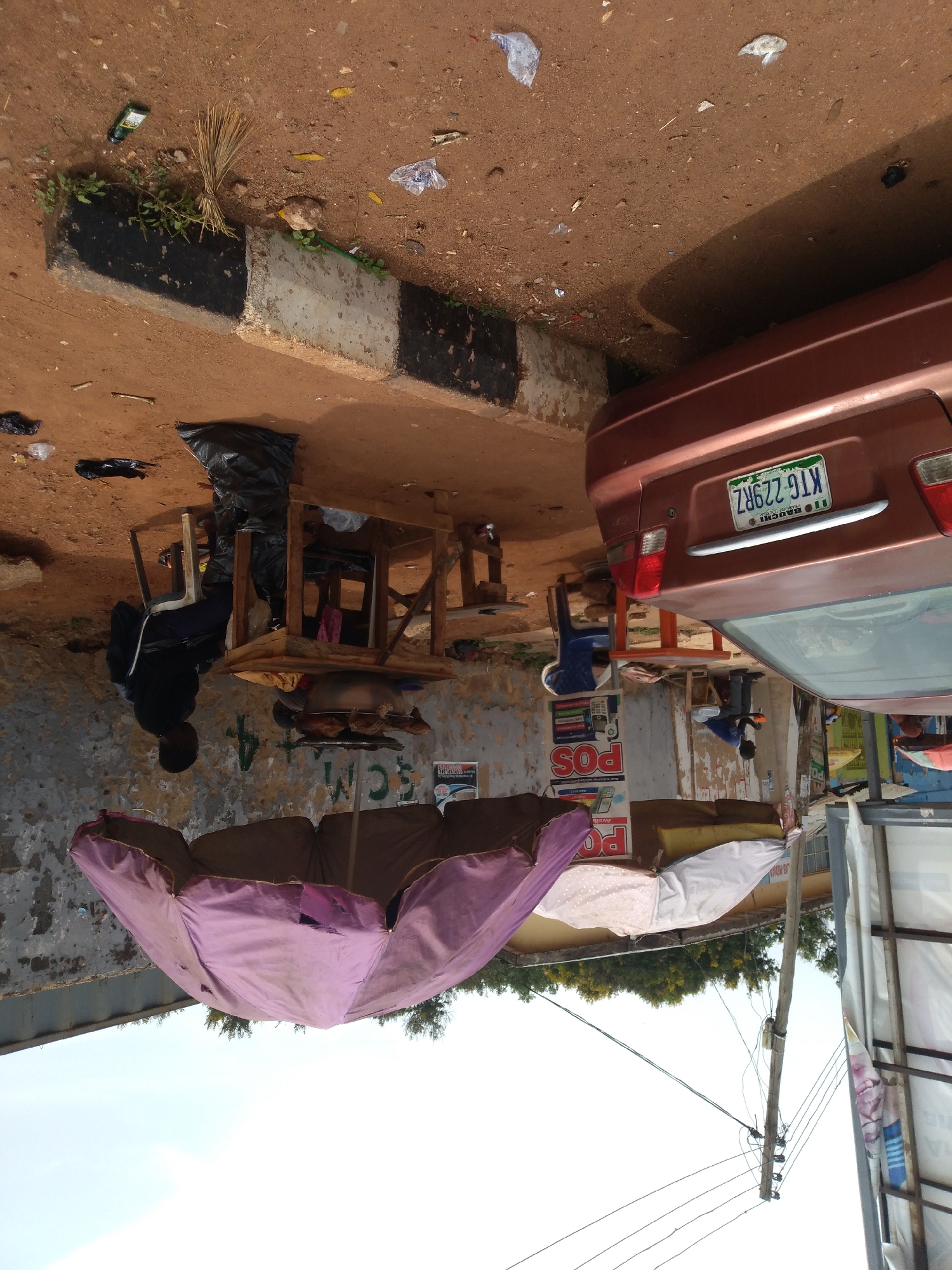 The image depicts an outdoor scene in Jos, Nigeria, featuring a roadside market stall. A person is seated at a wooden table, which is laden with goods, including a large pot. Behind the table, a black plastic bag is placed on the ground. A faded pink umbrella is propped open, serving as a canopy. In the background, the rear of a reddish-brown car is visible, displaying a license plate with the characters "KTG 229RZ". The ground is covered with reddish-brown earth, and scattered debris, including plastic bags and leaves, litters the area. The scene appears to be set along a street or pathway, with a concrete curb painted in black and white stripes. A wooden pole with electrical wires runs through the scene. The overall impression is one of a bustling, albeit somewhat unkempt, commercial area.