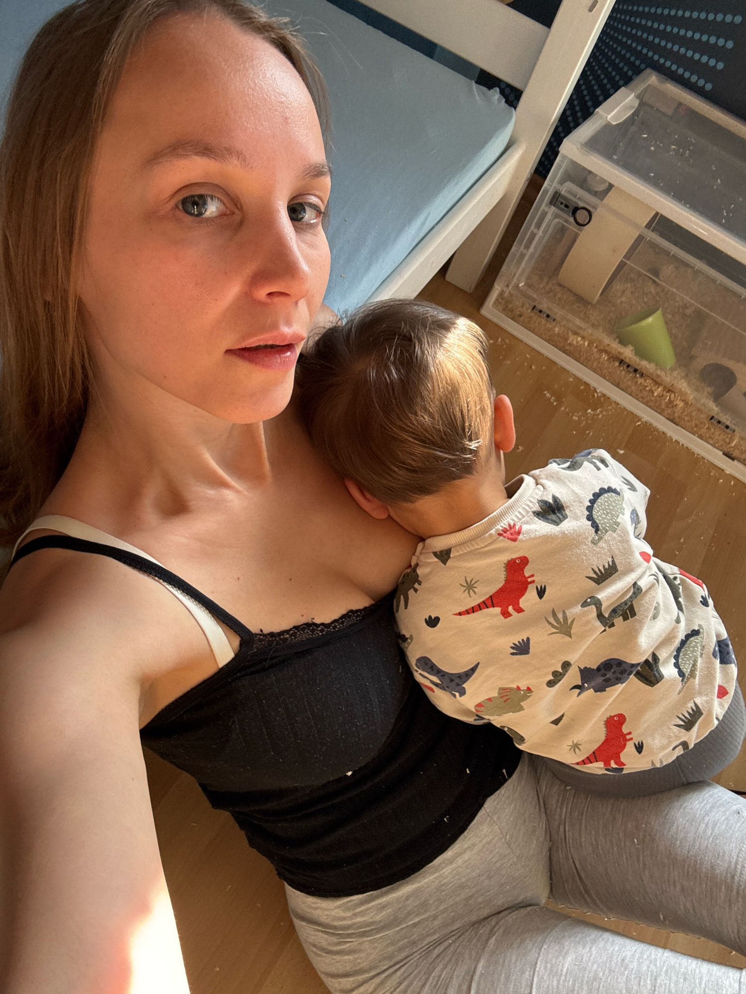 The media file depicts a close-up, overhead shot of a woman holding a baby. The woman, with blonde hair, is looking towards the camera with a neutral expression. She is wearing a black tank top with white straps and gray leggings. The baby is nestled against her chest, with their back to the camera. The baby is wearing a light-colored long-sleeved shirt with a dinosaur print.
The scene appears to be indoors, with a wooden floor visible. To the right of the subjects, there is a white bunk bed with a light blue sheet. Further to the right, there is a clear plastic container with bedding material and a small green cup, possibly an animal habitat. The lighting suggests it is daytime, with sunlight casting a warm glow on the woman's arm and chest. The overall mood is intimate and peaceful. No visible text is present in the image. The location context is Überherrn, Germany.