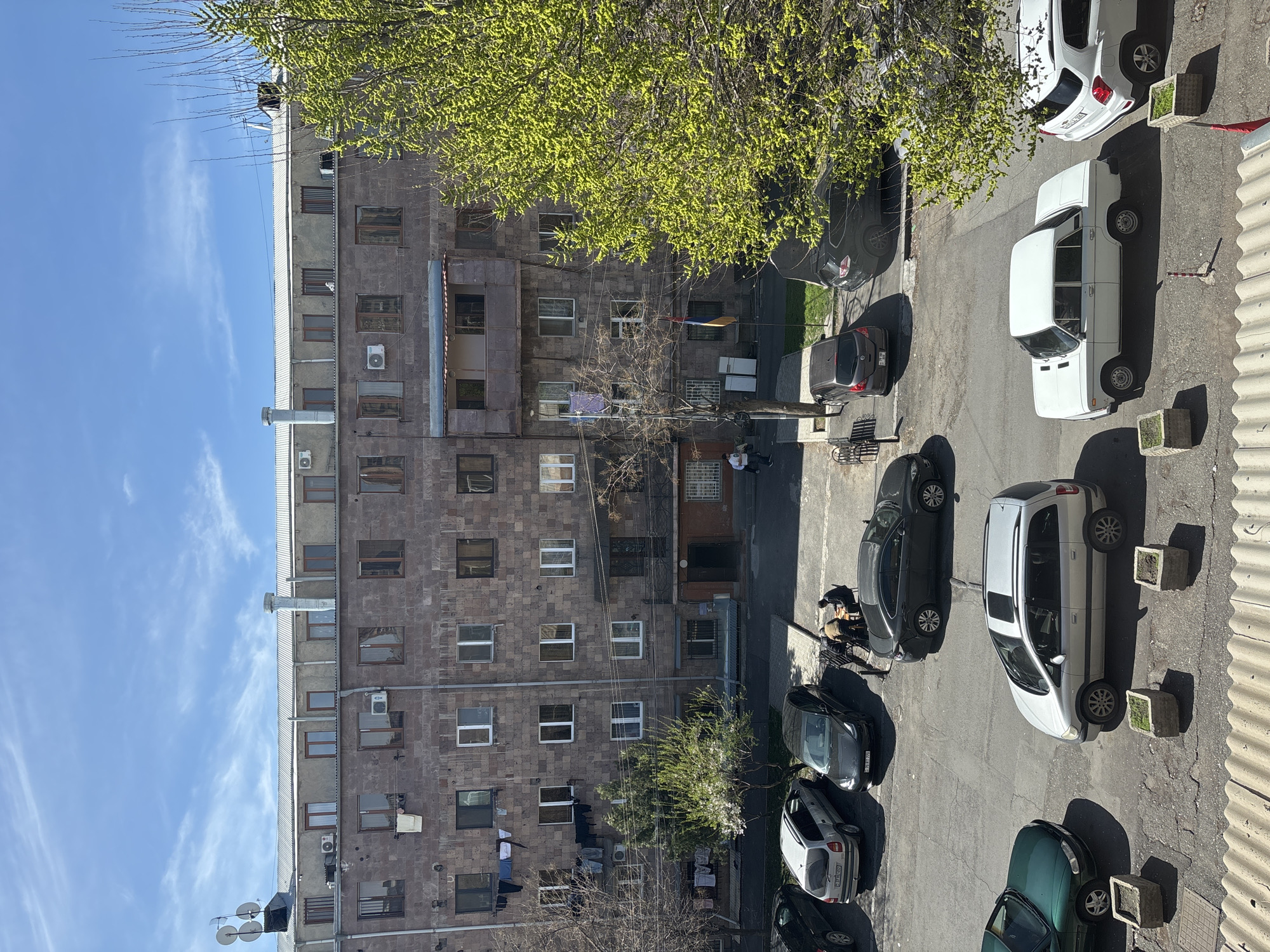 An elevated view depicts a Yerevan, Armenia urban scene. A multi-story building with a stone facade dominates the left. A variety of parked cars line the street. A tree with new green leaves is overhead. Laundry hangs from lines extending from the building. Two individuals appear to be working on something near a parked car. The sky is blue with scattered clouds.