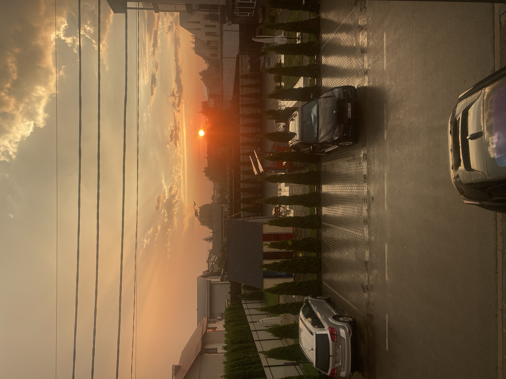 The image captures an outdoor scene during sunset in Novi Petrivtsi, Ukraine. The sun, a bright orange orb, is low on the horizon, casting a warm, golden light across the sky, illuminating scattered clouds. Power lines stretch horizontally across the upper portion of the frame, with buildings and trees visible in the background.
In the foreground, a paved area, possibly a parking lot or driveway, is wet, suggesting recent rain. Several cars are parked: a dark-colored sedan with license plate "MYD 781" is prominently featured, and a lighter-colored SUV with license plate "AK 6683 OX" is visible further down. The parked cars and the paved surface indicate a residential or commercial setting. The presence of manicured hedges and modern architecture suggests a well-maintained, possibly suburban or newly developed area. The overall mood is peaceful, with the soft light of the sunset creating a serene atmosphere. The time of day is clearly sunset, and the wet ground indicates recent precipitation.