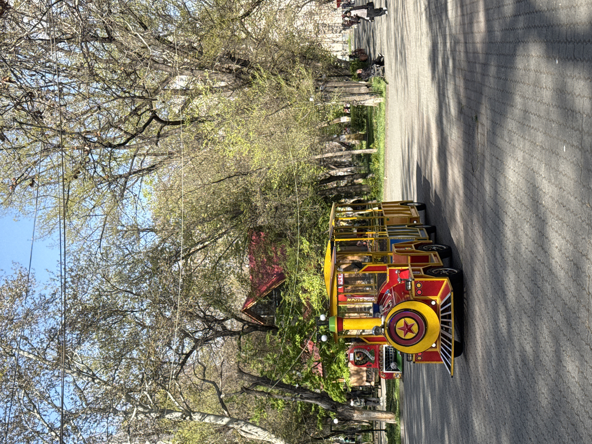 The image depicts a toy train, painted red and yellow, parked on a paved pathway in a park setting. The train, complete with a prominent star emblem on its front, appears to be a tourist attraction or amusement ride, with multiple passenger cars behind the locomotive. The path, patterned with rectangular pavers, stretches into the distance, leading past rows of trees with budding spring foliage. A few figures are visible further down the path, engaged in various activities. The environment is characterized by a mix of shade and sunlight dappling the ground. The location is Yerevan, Armenia.