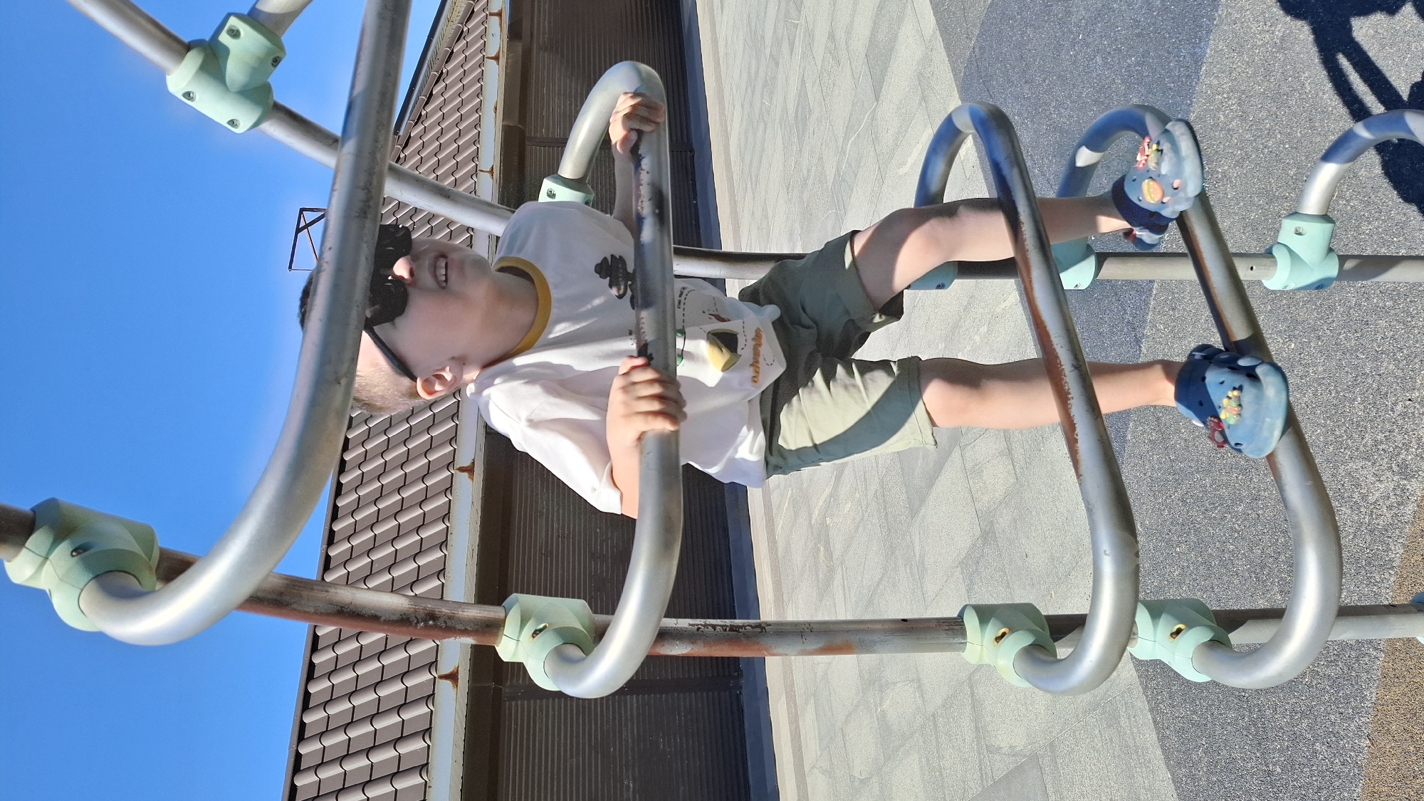A young child is depicted hanging upside down on a metal climbing structure under a bright blue sky. The child, likely a boy, is wearing dark sunglasses, a white short-sleeved t-shirt with a graphic that includes a cartoonish design and the faint word "nature" or similar, and olive green shorts. His feet are clad in light blue clogs adorned with colorful flower-like embellishments, which are hooked onto one of the metal bars. His hands grip other metal bars of the structure.
The child is smiling, indicating enjoyment and play. The climbing structure consists of silver-colored metal bars connected by light blue or teal plastic clamps. The background features a clear, deep blue sky on the left side, and a dark tiled roof of a building is visible on the right. The ground beneath the structure appears to be a paved surface, possibly asphalt or concrete. The scene is brightly lit by natural sunlight, suggesting a clear and sunny day, likely during daytime. The image captures a moment of active play and childlike wonder from a low-angle perspective, emphasizing the child's upside-down position.