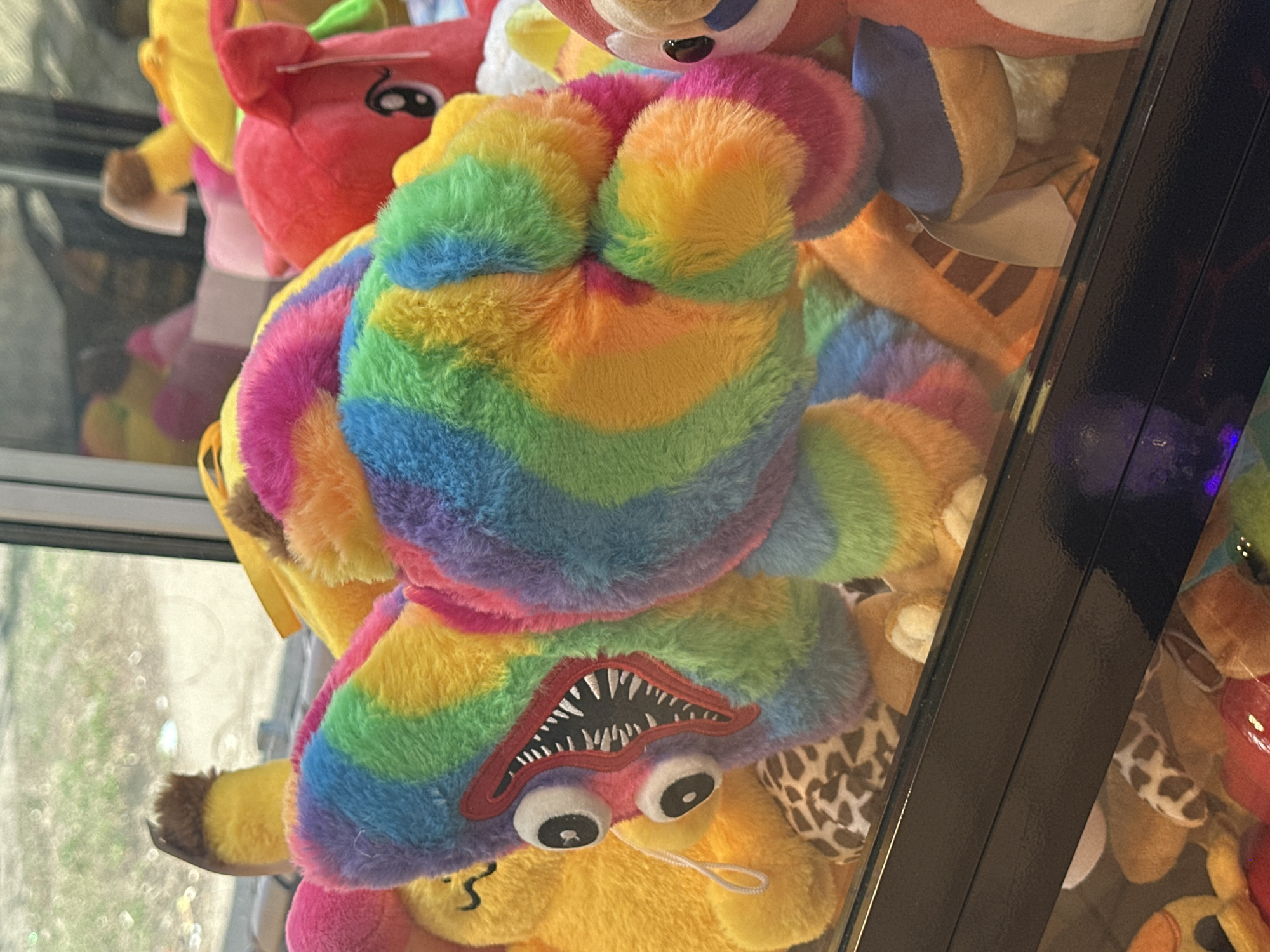 The image shows a claw machine filled with plush toys. The main subject is a multi-colored, rainbow-striped plush toy with a large, toothy mouth and prominent eyes, reminiscent of the character "Huggy Wuggy" from the game "Poppy Playtime." Other plush toys of various colors and shapes are visible in the background, including a red animal and some giraffe-print toys. The black frame of the claw machine is visible in the foreground. The scene is likely indoors and based on the toys it could possibly be at the shopping mall. Since there is no definitive evidence in the image, it can not be concluded that the location is Moscow, Russia.