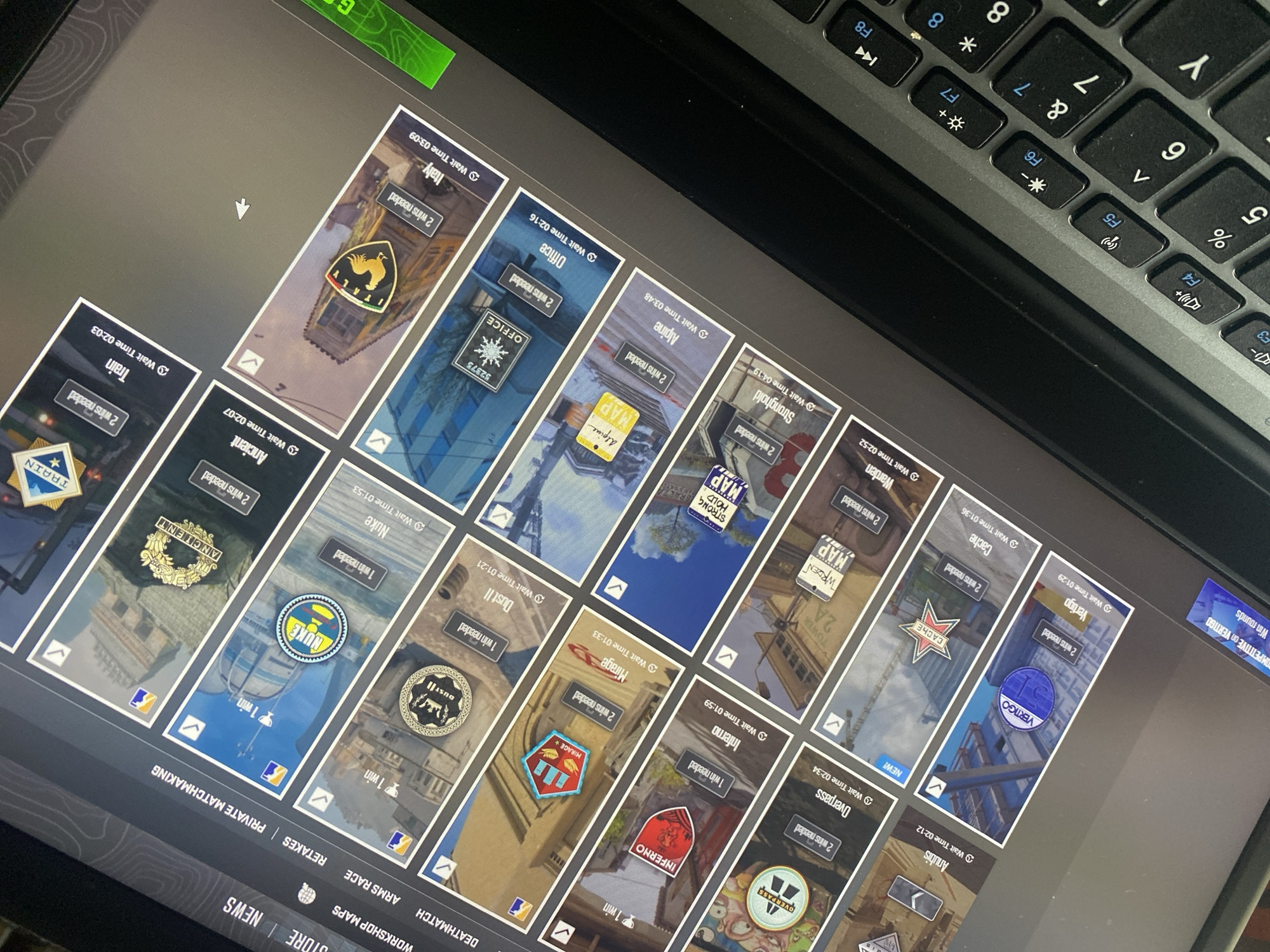 A computer screen displays a video game interface, likely a map selection screen, viewed from an elevated, angled perspective. The screen is filled with a grid of rectangular cards, each representing a different game map.
Each map card shows a small icon or image, the map's name, a "Wait Time" (ranging from around 1 to 4 minutes), and a requirement for either "1 win needed" or "2 wins needed." Visible map names include "Italy," "Office," "Alpine," "Stronghold," "Warden," "Cache," "Vertigo," "Anubis" (marked "NEW!"), "Overpass," "Inferno," "Mirage," "Dust II," "Nuke," "Ancient," and "Train." Map icons feature elements like a rooster for Italy, a snowflake for Office, an eagle for Ancient, and a train for Train. One card also shows "COMPETITIVE on VERTIGO" and "Win rounds."
At the bottom of the screen, several menu options are visible, including "STORE," "NEWS," "WORKSHOP MAPS," "DEATHMATCH," "ARMS RACE," "RETAKES," and "PRIVATE MATCHMAKING."
A black and grey laptop keyboard is partially visible in the top right corner of the image, showing function keys F3 through F8, and number keys 5 through 8. Many keys display secondary functions in blue. The screen interface has a dark, patterned background with a prominent green bar at the top left.