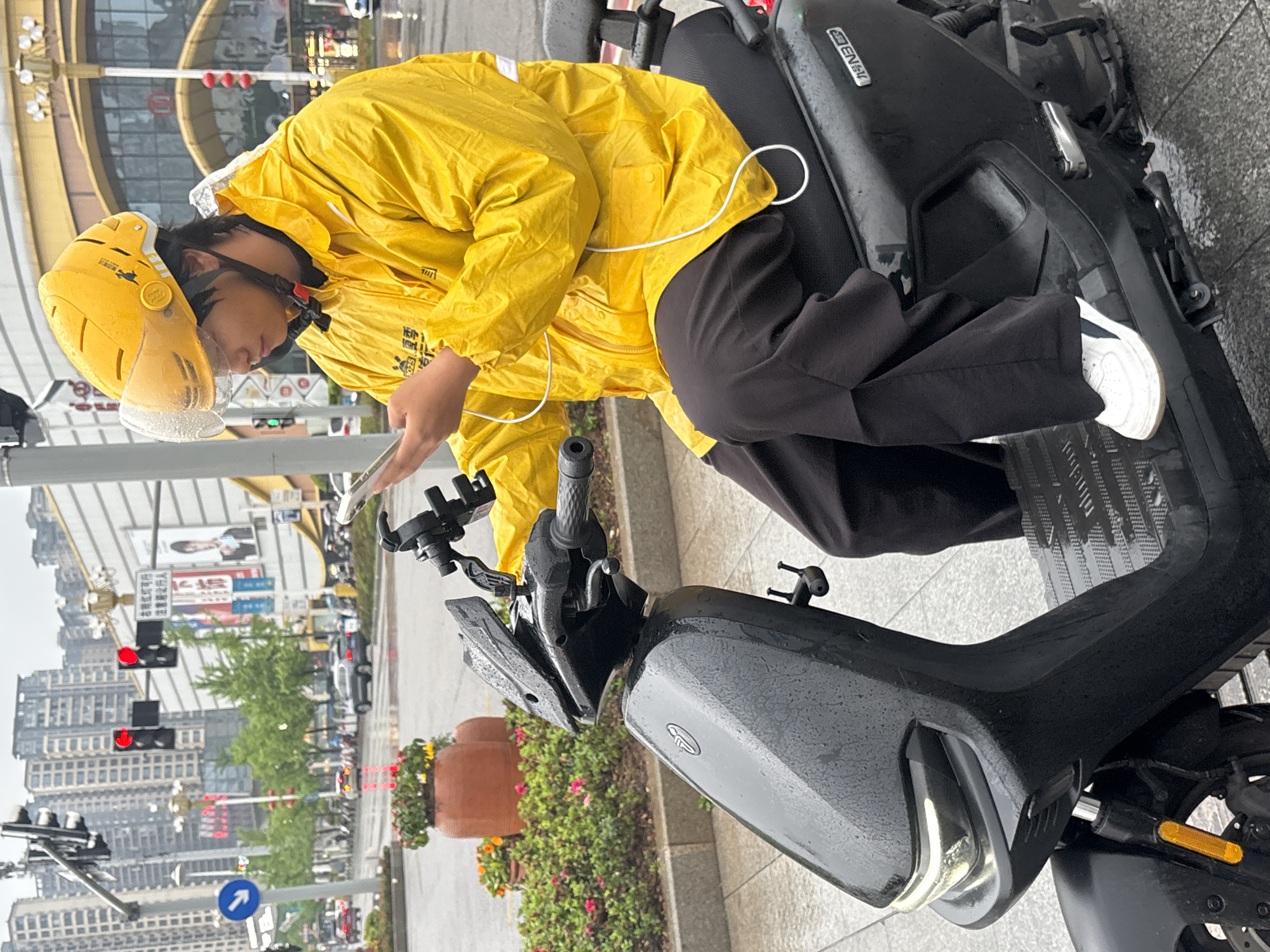 A person in a yellow raincoat and matching helmet is seated on a black scooter on a city street. The person appears to be using a mobile phone mounted on the scooter's handlebars. The scooter itself is damp with visible water droplets.
In the background, there is a cityscape with high-rise buildings, traffic lights (showing red), and various street signs, including Chinese characters and logos. The plants and flower pots line the street.
The person is wearing black pants and white sneakers. The scooter appears to be a 'Ninebot' model, based on the logo on the vehicle.
Additional Chinese text on a yellow placard on the scooter translates to "Micro red light is feasible, pay attention to giving way to pedestrians, 1025".
Based on the characters "Jiangjin" appearing in the Chinese text, the city is Jiangjin, China.