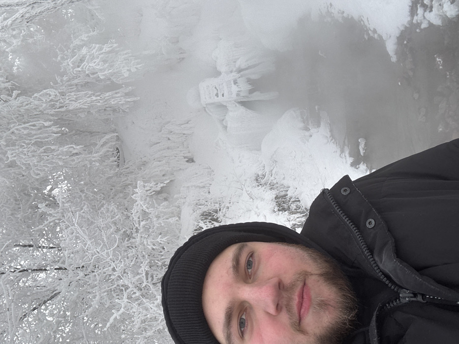 A male individual is depicted in a selfie-style photograph within a winter landscape. The subject, positioned on the left, has light skin, blue eyes, and a light brown beard, wearing a black beanie and a black hooded winter jacket. His cheeks appear ruddy from cold exposure. The background consists of trees and branches heavily coated in white rime ice, creating a frosted, crystalline appearance. To the right of the subject, a body of open water, likely a stream, emits significant steam or mist, indicating a warmer water temperature amidst cold ambient air. The edges of the water body are partially frozen. Further in the background, a man-made structure, possibly a wooden ladder or grate, is visible, entirely encased in thick ice and icicles, situated within snow and ice formations near the water's source. This scene is identifiable as the Warm Spring (Теплый источник) located in Krasnoyarsk, Russia, known for its steaming water and heavy rime ice formation in winter.