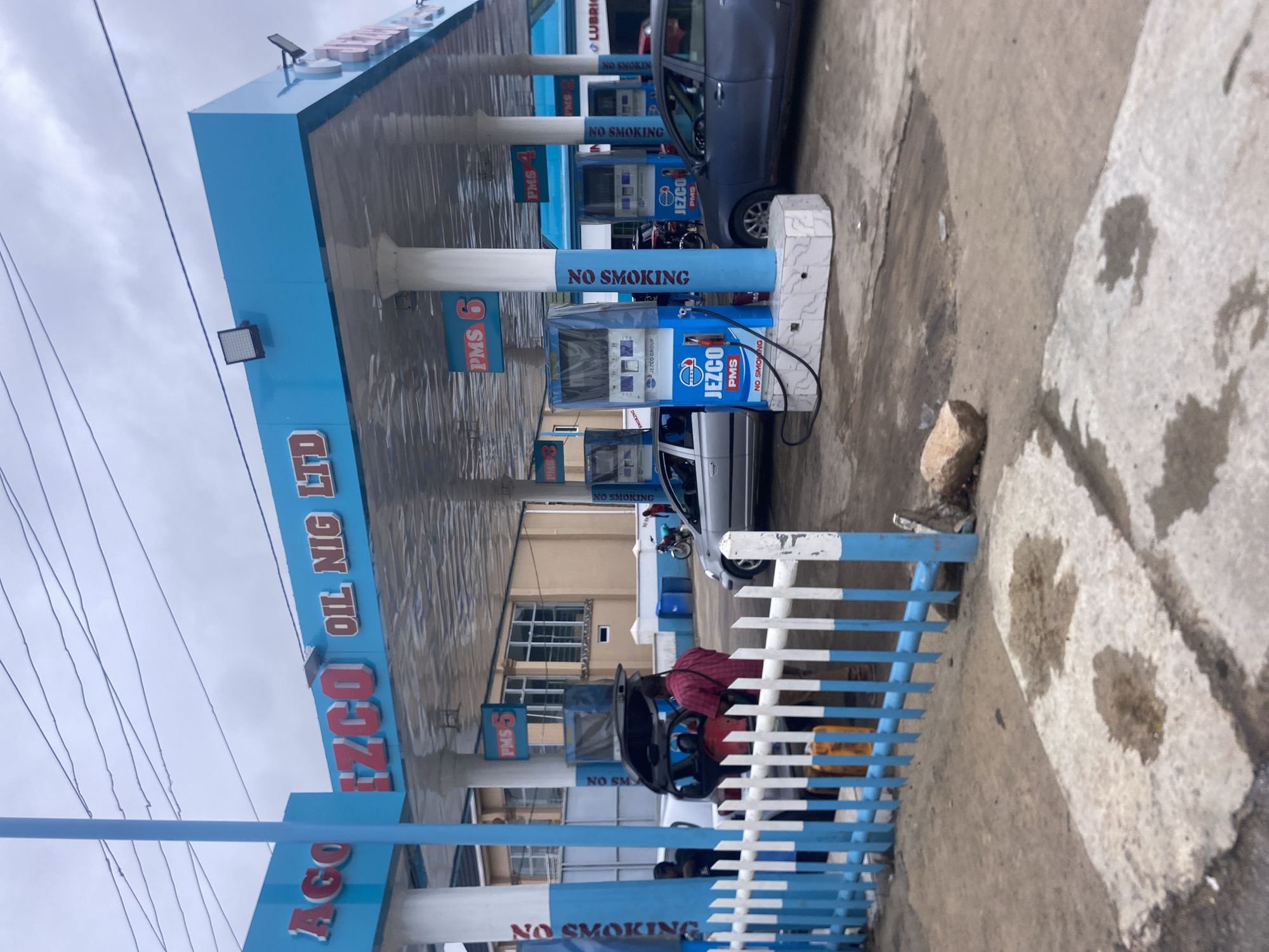 This is a photograph of the Jezco Oil Nig Ltd. gas station in Jalingo, Nigeria. The gas station has a blue canopy supported by white pillars. The Jezco logo is visible on the canopy and on the gas pumps. There are several cars at the gas station, including a silver sedan and a dark blue sedan. A man in a red shirt is standing by the open trunk of a car, and other people are also present. Some gas pumps have "PMS 6", "PMS 5", "PMS 4", and "PMS 3" labels indicating different fuel grades or pump numbers. Several "NO SMOKING" signs are displayed. The ground is paved with concrete and asphalt, with some puddles of liquid. A white and blue striped fence is in the foreground. A motorcycle is parked near one of the gas pumps. The sky is cloudy.