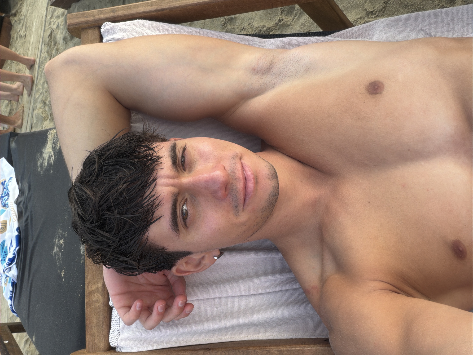 A young man lies on a lounge chair on a sandy beach, his head resting on his arm. He has dark, messy hair and is shirtless. His gaze is directed towards the camera. In the background, several pairs of legs are visible, suggesting other people are nearby. The scene is set on a beach, likely in Stavros, Greece, given the context.