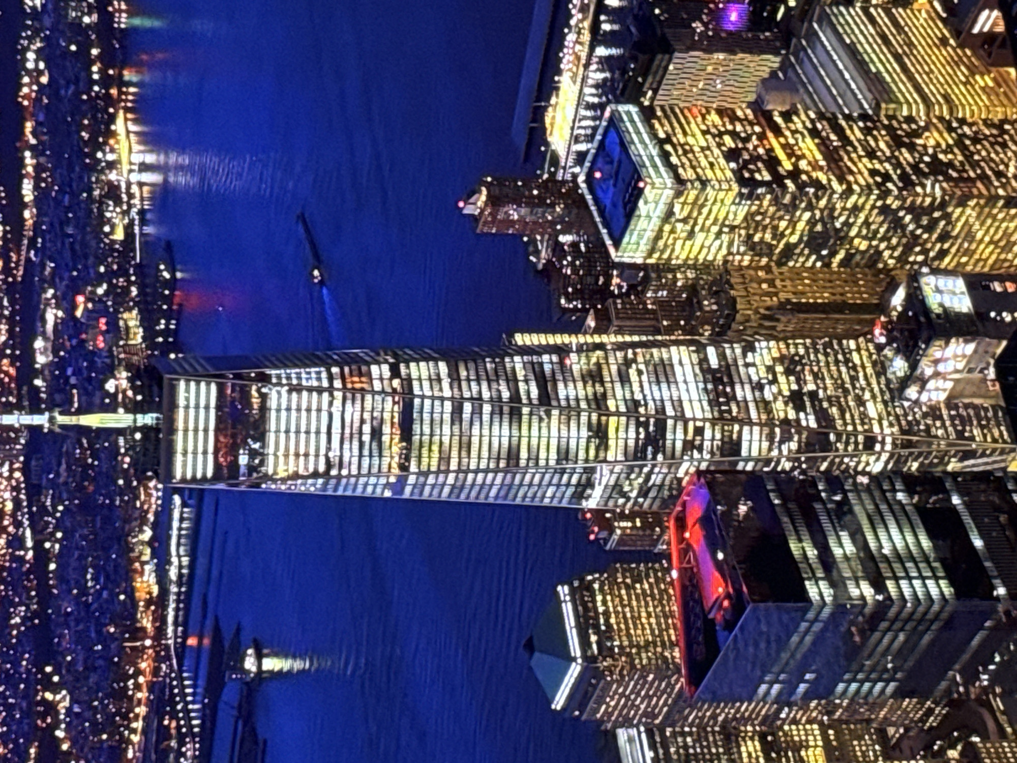 The image shows a nighttime aerial view of a densely built urban landscape dominated by skyscrapers illuminated with countless lights. The central and largest building is a modern skyscraper with a reflective facade, its windows glowing with warm light, suggesting occupancy. This building tapers towards its summit, which is topped by a spire or antenna.
Surrounding the central skyscraper are other tall buildings of varying architectural styles, also lit from within, creating a dazzling display of light against the dark sky. Some of these buildings have unique features; one in the foreground on the left has a dark, faceted facade topped with red lights, while another to its right is a square-shaped building with a bright blue illuminated roof.
In the background, a large body of dark water, likely a harbor or river, reflects the lights of the city. Several vessels, including what appears to be a barge or cargo ship with a lit wake, are visible on the water. The distant shoreline is densely populated with more buildings and lights, indicating a sprawling metropolis. The overall scene is one of vibrant urban activity at night.
This image is not of Burgas, Bulgaria. It depicts the skyline of a major city, likely New York City, given the prominent presence of a skyscraper resembling One World Trade Center and the surrounding architecture.