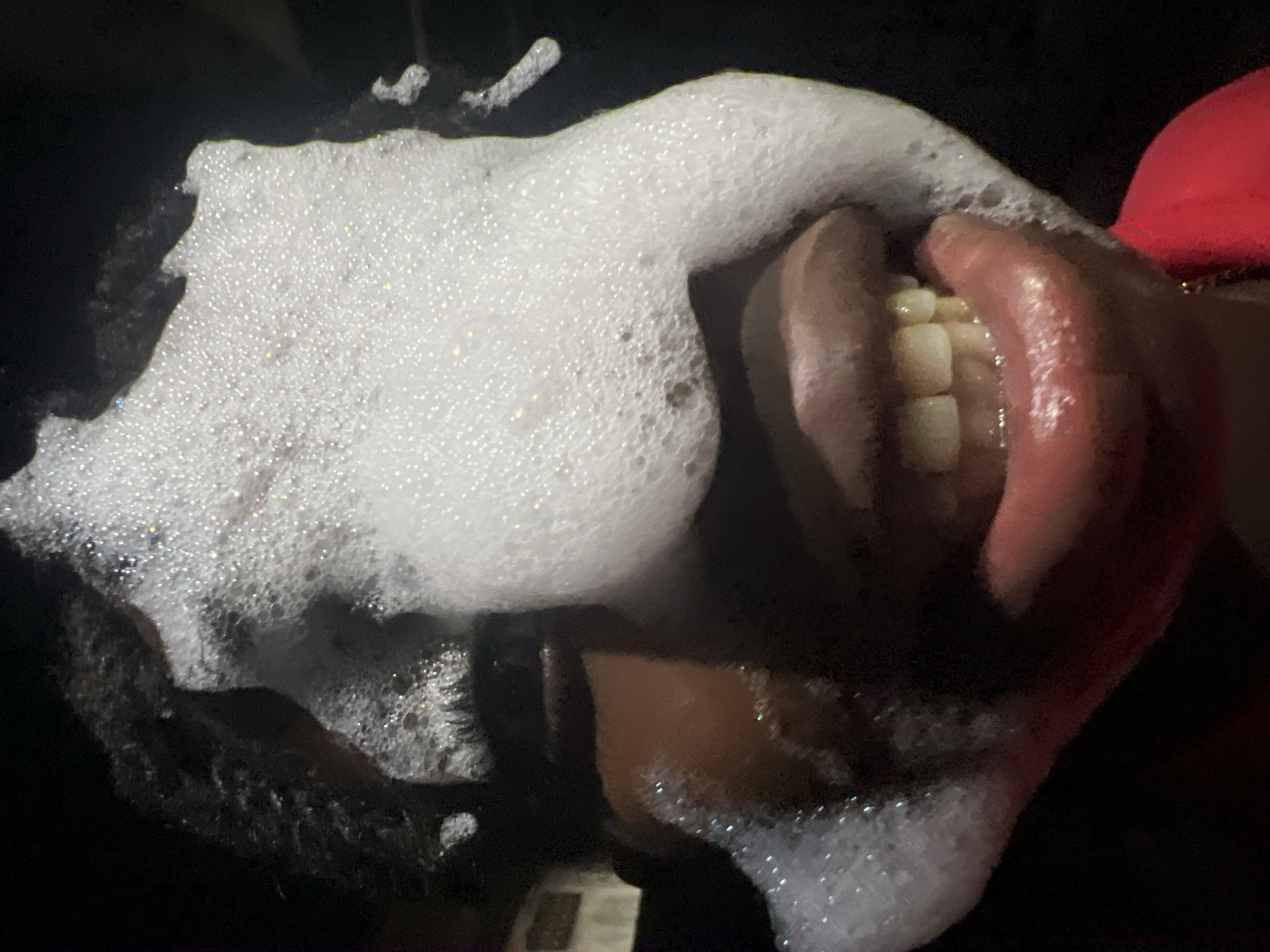 This is a close-up, eye-level, outdoor photograph of a person's face covered in white foam. The foam obscures much of the face, including the eyes and forehead, but the mouth and chin are visible. The person's lips are parted, revealing their teeth, and they appear to be smiling. The foam is textured and bubbly, with some areas appearing more solid than others. The background is dark and out of focus, suggesting the photograph was taken at night or in a dimly lit environment. The location is specified as Nkpor, Nigeria.