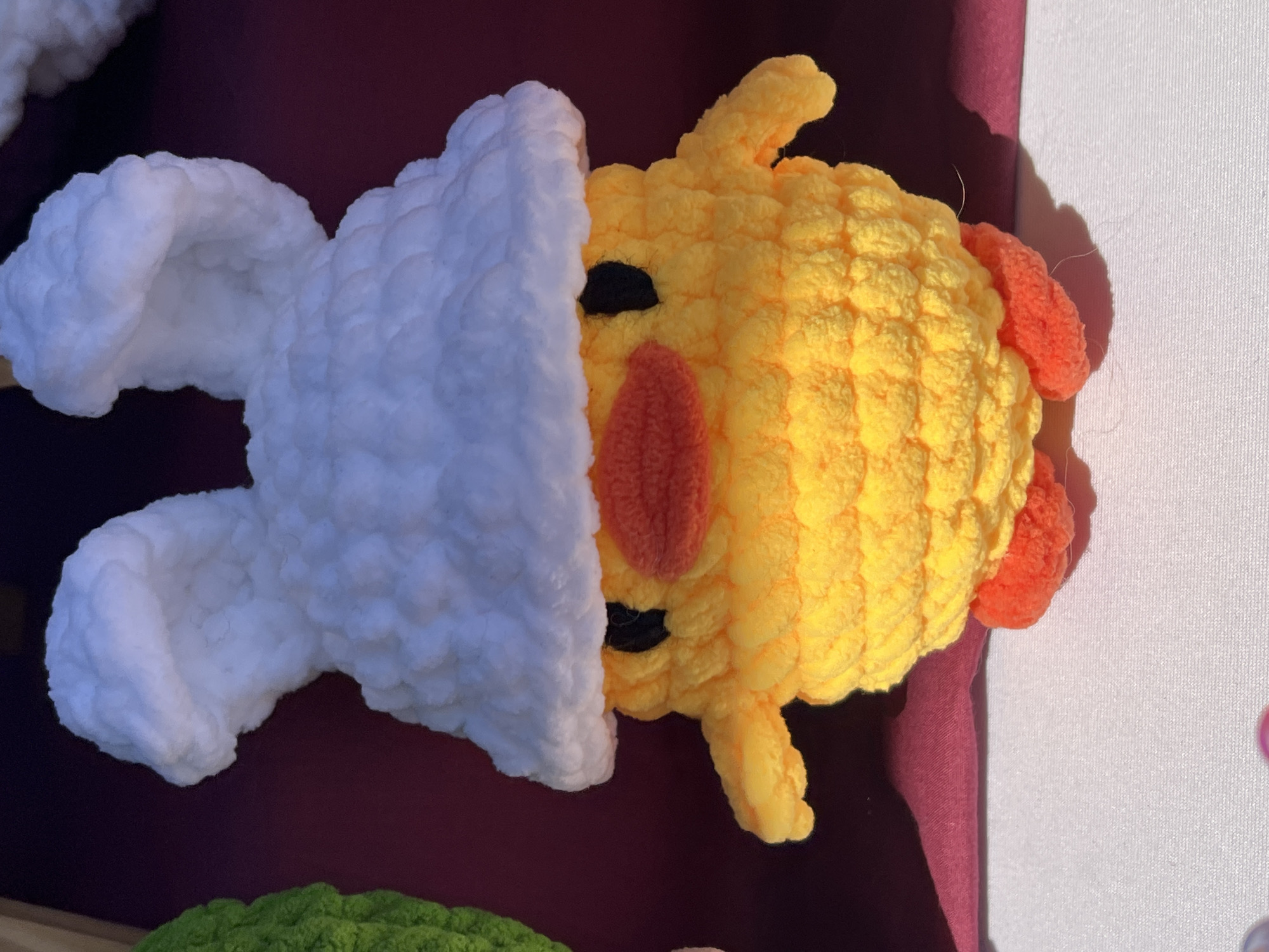 A close-up, eye-level shot shows a handmade crocheted or knitted chick wearing a rabbit costume. The chick is yellow with orange features: a small beak and wings. It has black eyes. The rabbit costume is white and covers part of the chick's head and body, with the rabbit ears visible to the left. The scene is set outdoors, likely at a crafts market or fair. The background includes a burgundy-colored material under the toy and a light-colored surface nearby. A small part of another crocheted item, colored green, can be seen in the lower-left corner. The lighting indicates a sunny day, casting shadows near the chick. The location of the image is unknown, therefore it cannot be determined the city of Arica, Chile.