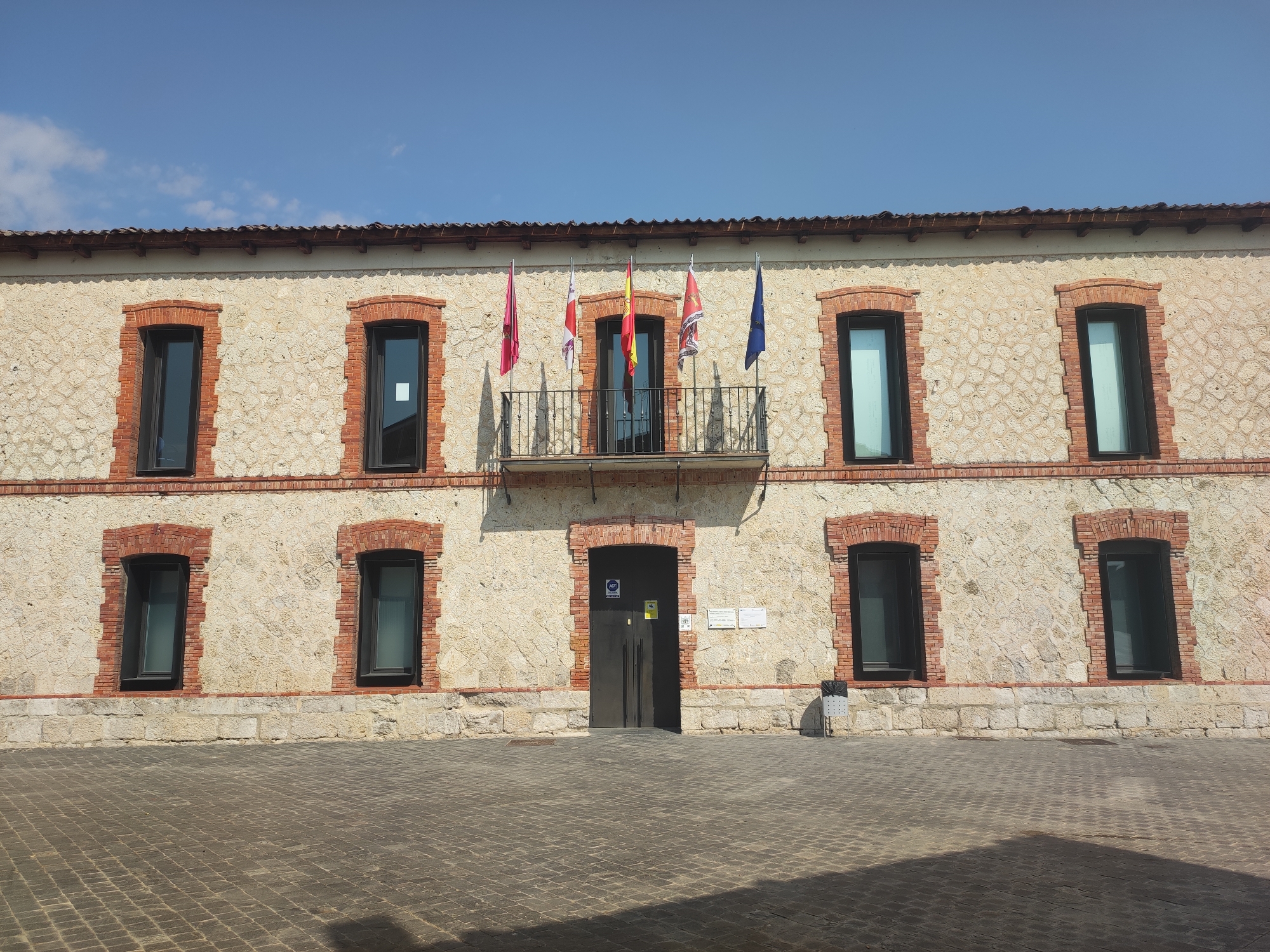 The image shows the exterior of a two-story building with a stone facade and red brick accents. The building has a row of windows on the upper level, with a balcony and several flags displayed above a central entrance. The lower level also has windows and the main entrance. The ground in front of the building is paved with cobblestones. The sky above is clear and blue. The building is identified as the Traspinedo Town Hall in Traspinedo, Spain.