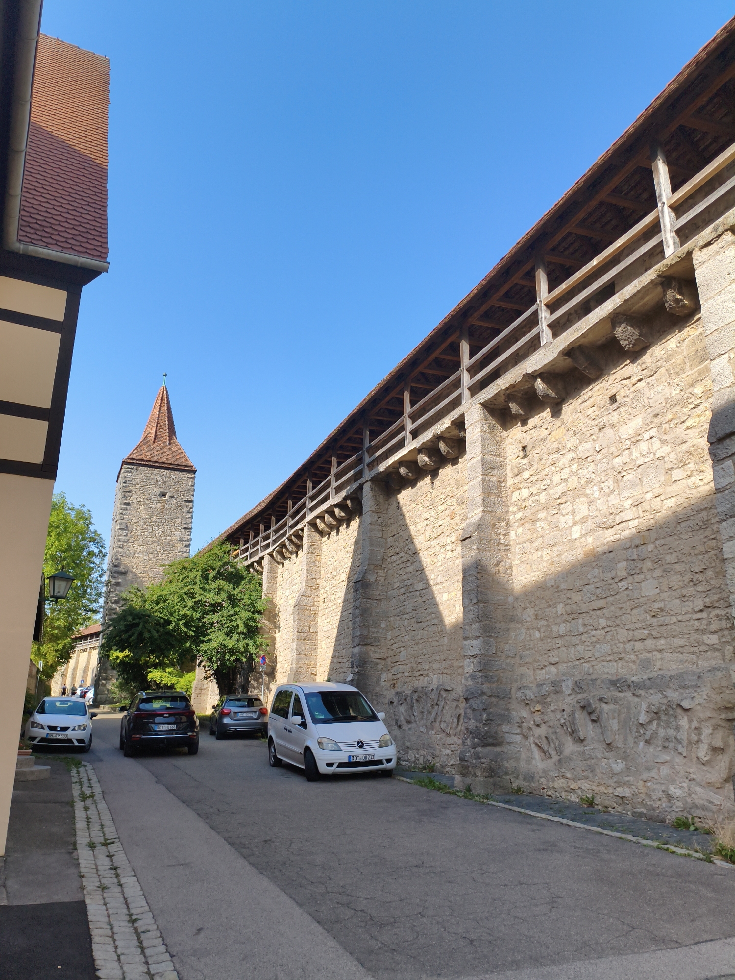 This image shows a street in Rothenburg ob der Tauber, Germany, with a prominent medieval wall and a tower. The street is paved with asphalt and has a cobblestone sidewalk on the left. Several cars are parked along the street. On the left, a building with a red-tiled roof and a white facade with dark beams is visible. To the right of the building, a tall stone tower with a red-tiled conical roof rises against a clear blue sky. Behind the tower, a long, high stone wall extends into the distance, topped by a wooden walkway with a red roof. The wall has a textured surface and appears to be an ancient fortification. The sunlight casts shadows across the wall and the street, indicating it is daytime. There are no people clearly visible in the foreground.
