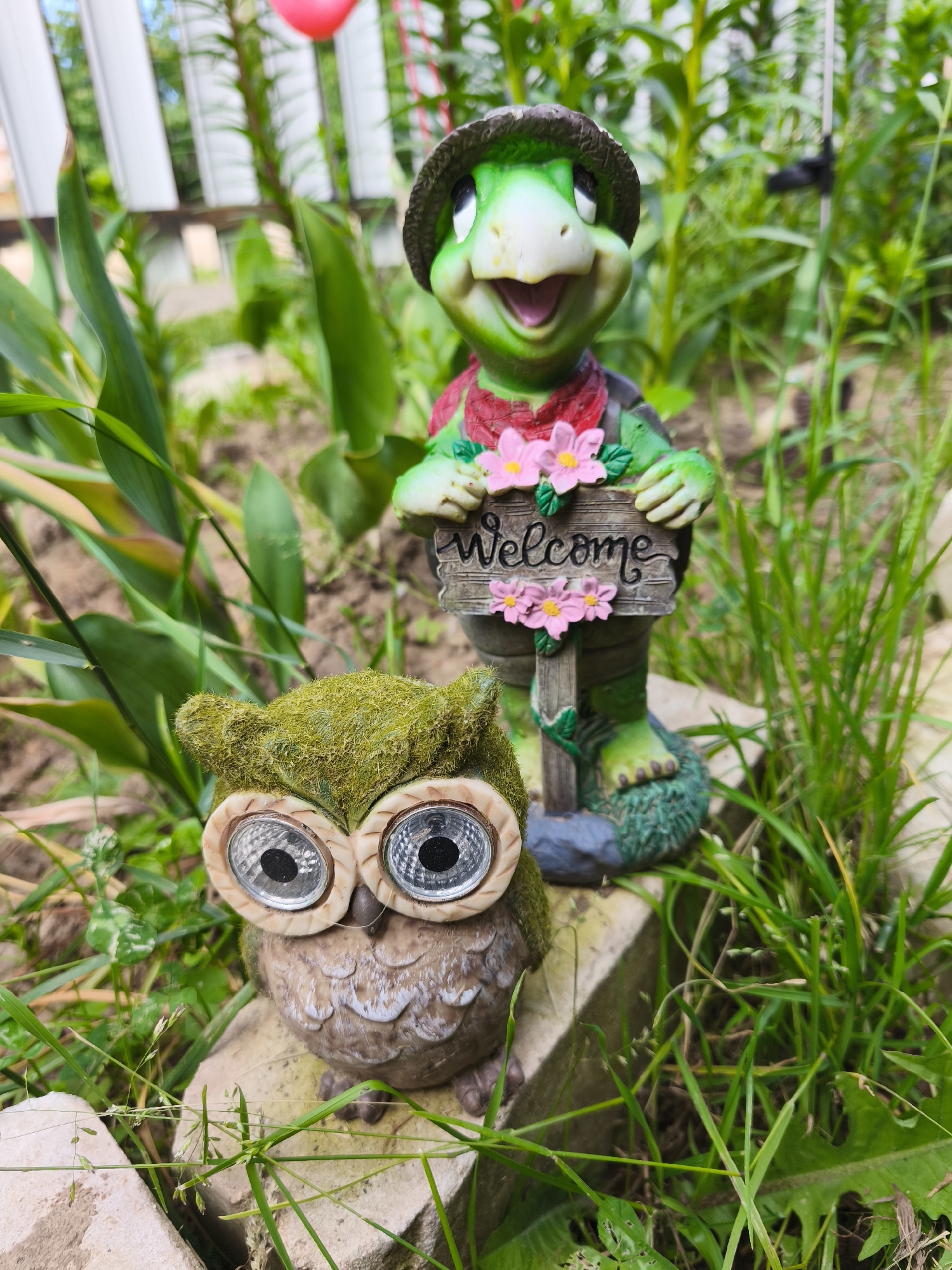 In a garden setting, two decorative figurines are positioned outdoors amongst green foliage and grass. In the foreground, a moss-covered owl figurine with large, reflective eyes sits on a light-colored stone. Behind and slightly to the right of the owl is a green turtle figurine wearing a hat and a red bandana. The turtle holds a wooden sign that reads "Welcome," adorned with pink flowers. The background shows a blurred white fence and more greenery. The scene appears to be in the city of Puchezh, Russia, given the context.