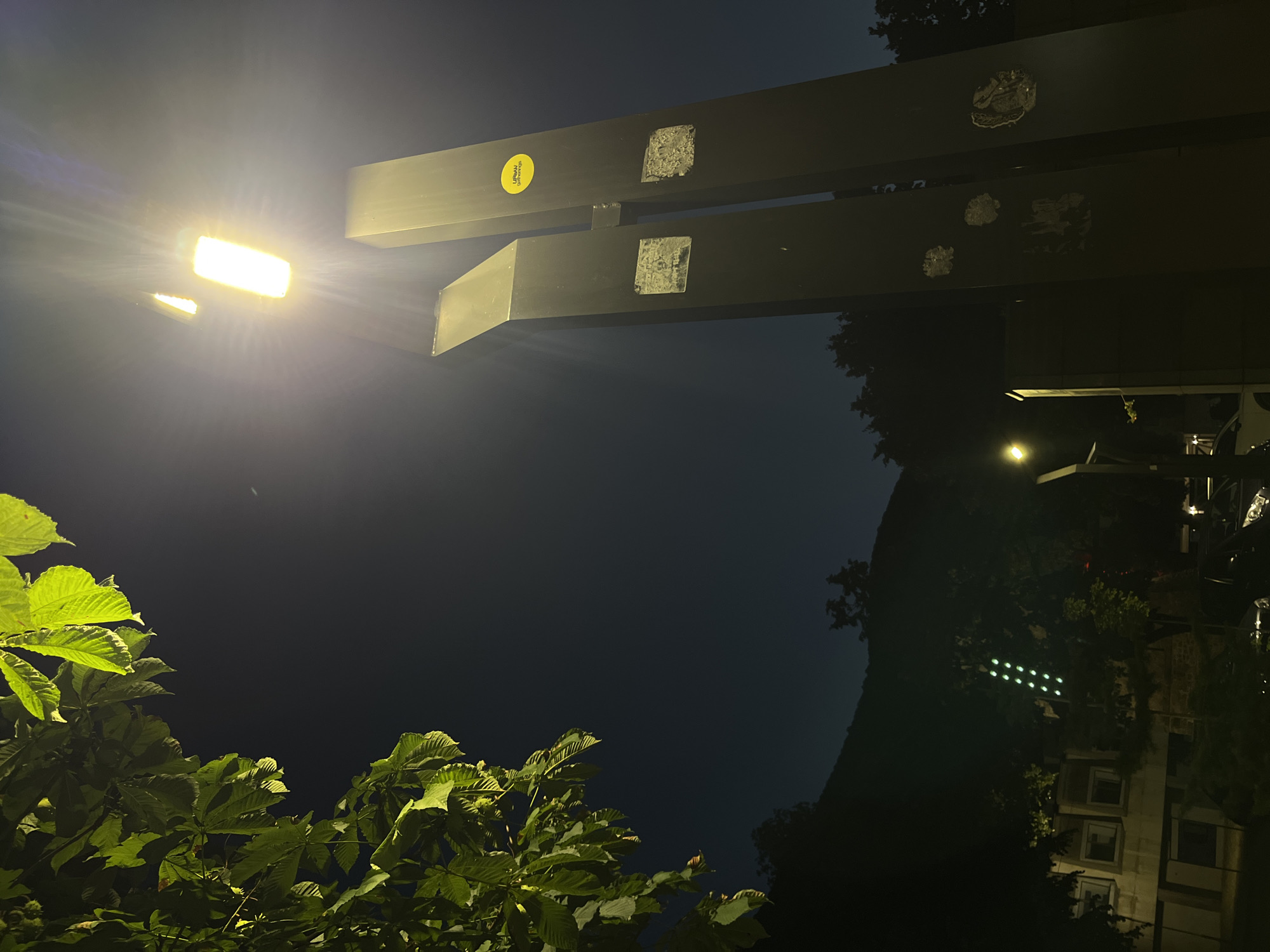 The image depicts a nighttime scene in Vratsa, Bulgaria, featuring a modern monument with two tall, dark vertical pillars. A bright streetlight illuminates the top of the monument, casting a strong glow. On the left side of the frame, lush green leaves from a tree hang down, their edges highlighted by the artificial light. In the background, the silhouette of a hill is visible against the dark sky. To the left, a building with lit windows and a traffic light displaying green lights are present. The overall impression is of a contemporary urban setting at night.