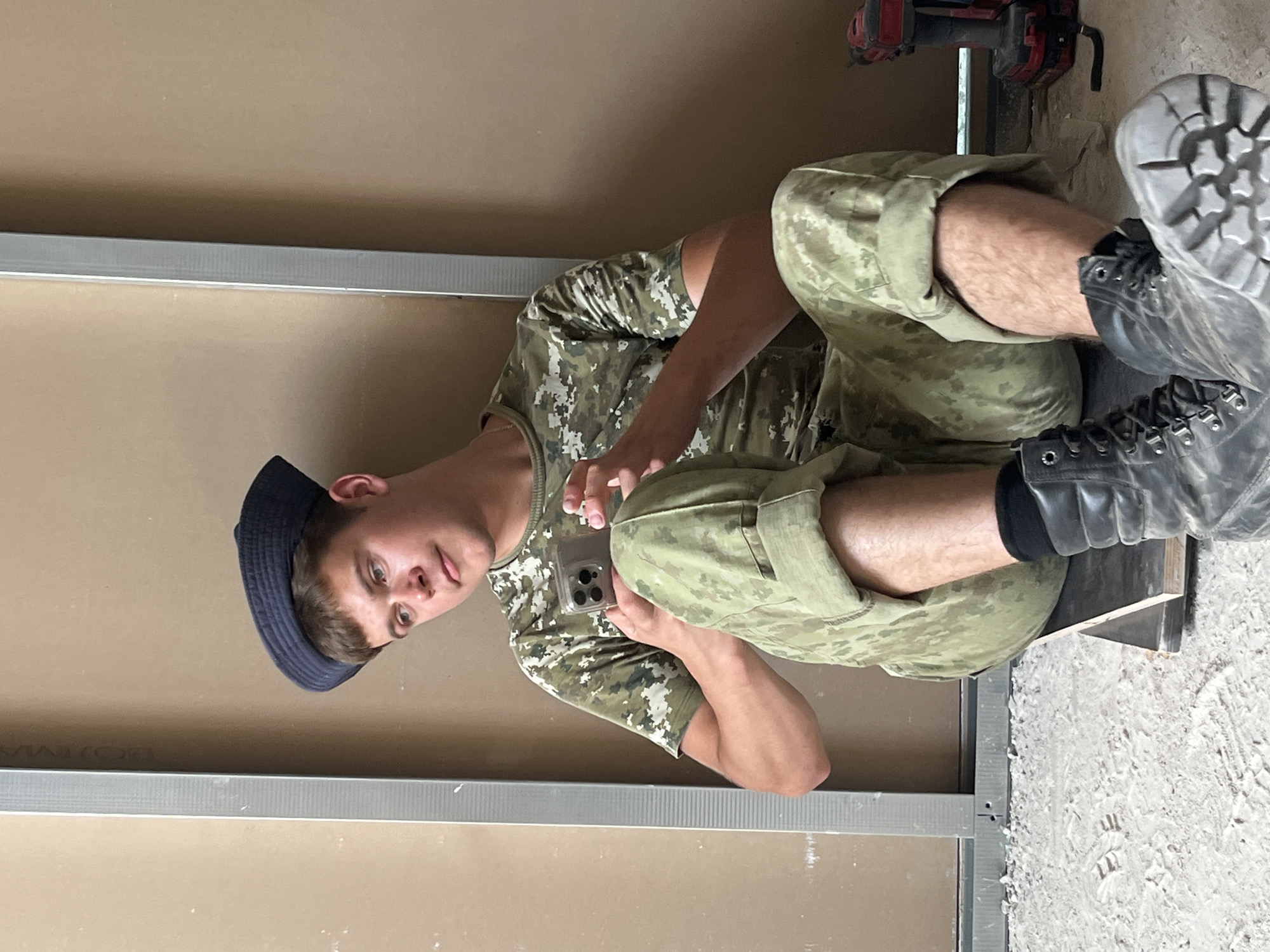 A young man in camouflage clothing sits on a wooden plank on a construction site. He wears a dark blue hat and black boots. He is holding a smartphone in his hands, looking at its screen. The background shows construction materials and tools, including a drill. The scene appears to be indoors, possibly in a building under construction, with drywall and metal framing visible. The lighting suggests it is daytime. The man's expression is neutral. There is no visible text in the image.