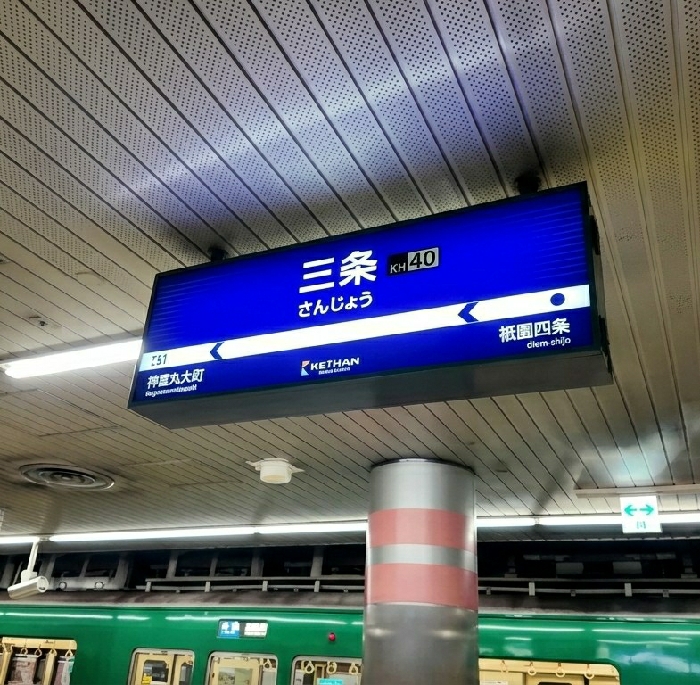 The image displays an interior view of an underground train station platform, identified as Sanjo Station (KH40) on the Keihan Main Line in Kyoto, Japan. A prominent, illuminated blue sign hangs from the ceiling, displaying "三条" (Sanjo) in Japanese characters and hiragana, along with the station code KH40. The sign also features the KETHAN (Keihan) logo and directional arrows indicating Jingumarutamachi (T51) to the left and Gion-Shijo to the right. The ceiling consists of light-colored, perforated acoustic panels arranged in parallel strips, equipped with fluorescent lighting fixtures. In the foreground, a large, cylindrical, silver-grey pillar with two pinkish-red horizontal bands partially obstructs the view. The side of a green train with white trim is visible on the track below, showing multiple windows and a door. An illuminated green exit sign is present in the background to the right of the pillar. No individuals are visible in the scene.