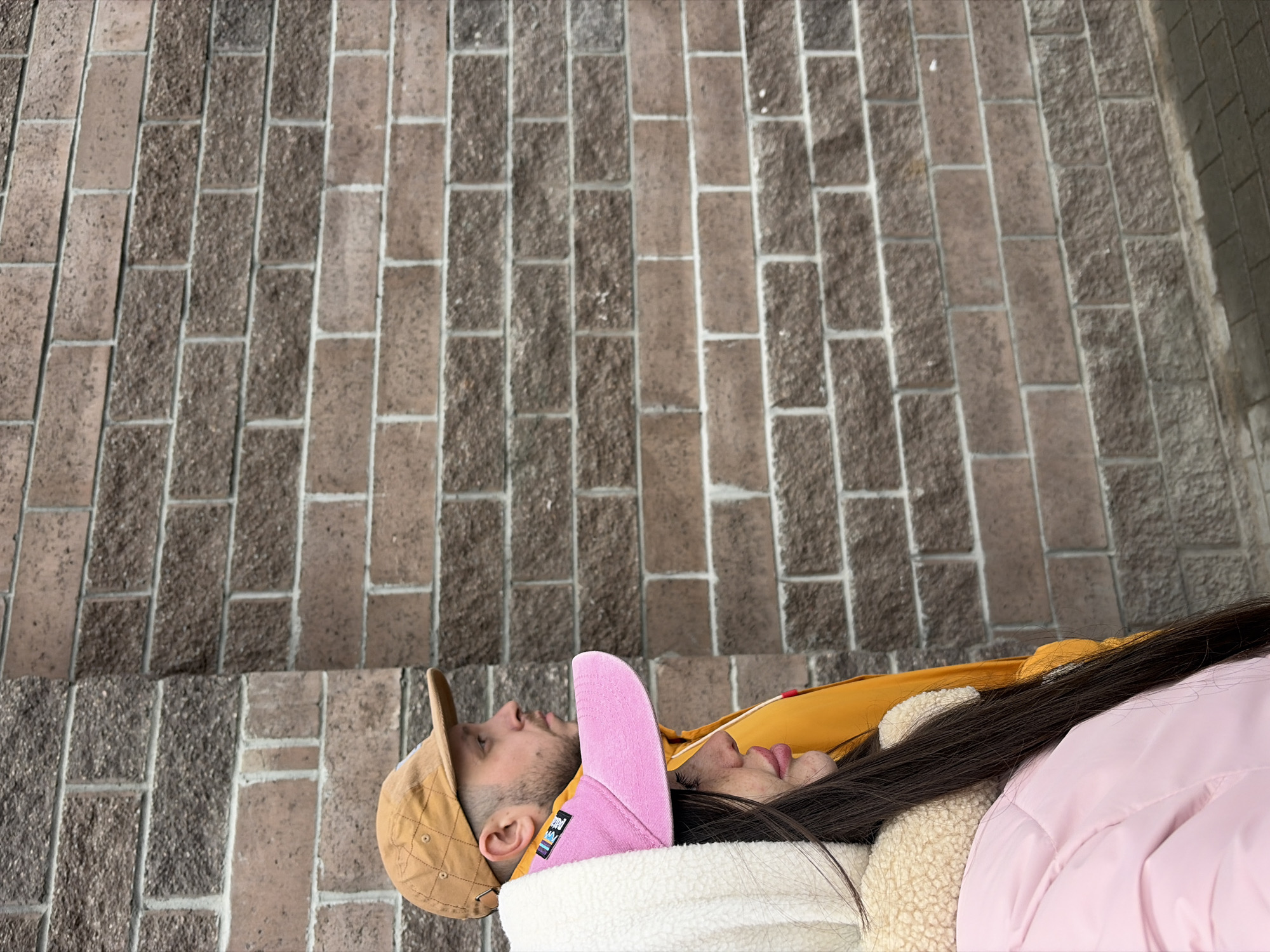 A medium-shot image shows two people lying side by side on a brick surface. The brick surface is composed of rectangular bricks in alternating shades of brown and gray, with light-colored grout between them. The bricks are laid in a running bond pattern.
On the left, a person wearing a tan baseball cap is lying on their back, with their head resting on a white, possibly fur-lined, surface. Next to them, another person is wearing a pink baseball cap and a pink jacket. The image is rotated to the left; the people are lying near a street, but the location cannot be determined from the image.