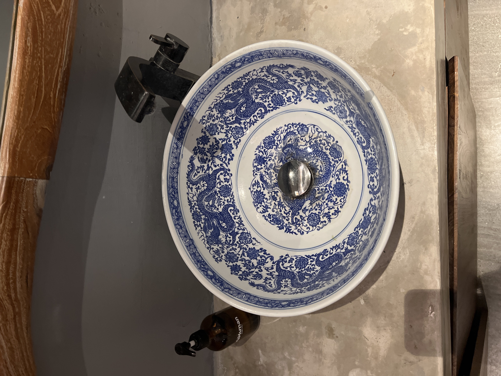 A top-down shot reveals a blue and white patterned washbasin, likely porcelain, mounted on a concrete countertop in a bathroom. The basin has a central drain and detailed designs of dragons and floral patterns. A black faucet is positioned to the left of the basin. Below and to the right of the sink, a brown bottle with a pump dispenser is visible. The background includes a wooden element and a grey wall. The location is North Kuta, Indonesia.