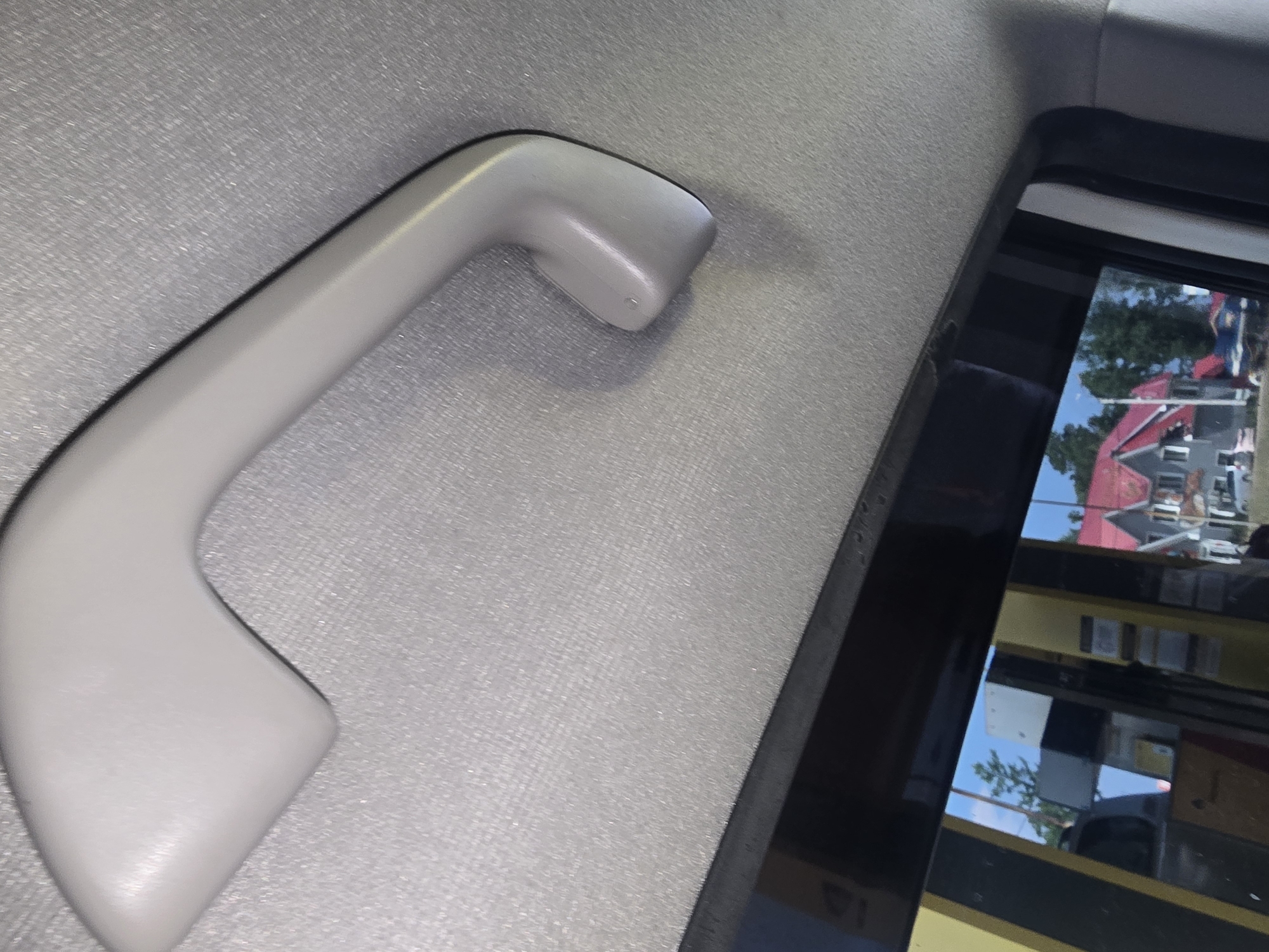 A grey interior grab handle is visible in the upper portion of the image, attached to a textured grey ceiling. Below the handle, a black window frame runs across the lower part of the image. Reflected in the window is a scene of a gas station with yellow and black pumps, and in the background, buildings with red roofs and green trees are visible under a blue sky. The location is identified as Turka, Russia. There are no people or animals present in the image.