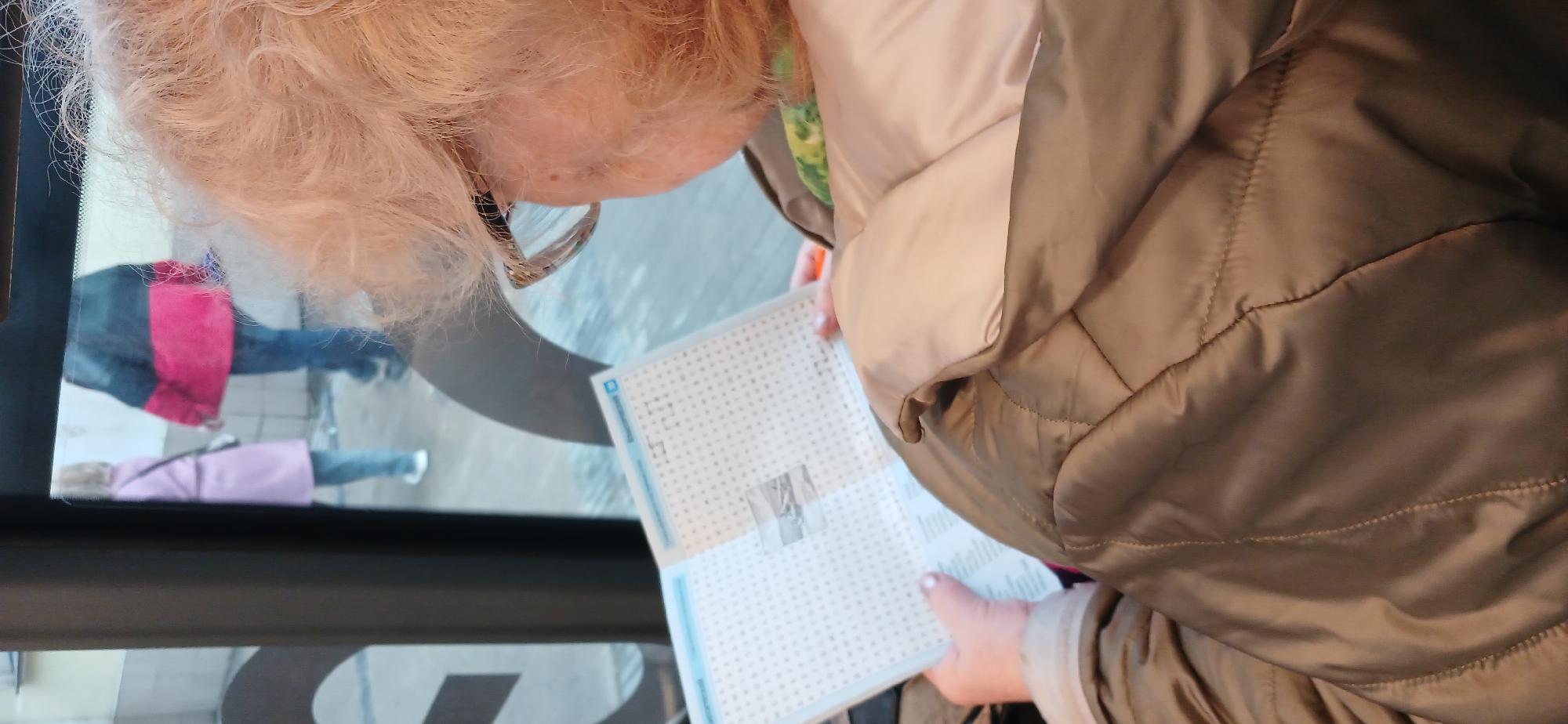 The image shows an elderly woman, likely in her 70s or 80s, with short, curly blonde hair, peering at a document filled with a grid pattern, possibly a puzzle or form. She is wearing glasses with a gold frame. She is wearing a puffy, light-colored, quilted jacket, with light brown/taupe coloring. She is inside a vehicle, presumably a bus or tram, as suggested by the presence of a window. The reflection in the window shows other people. Based on the presence of a blue stripe on the form and a blurred logo, it may be related to the public transportation system. This image may have been taken in Moscow, Russia.