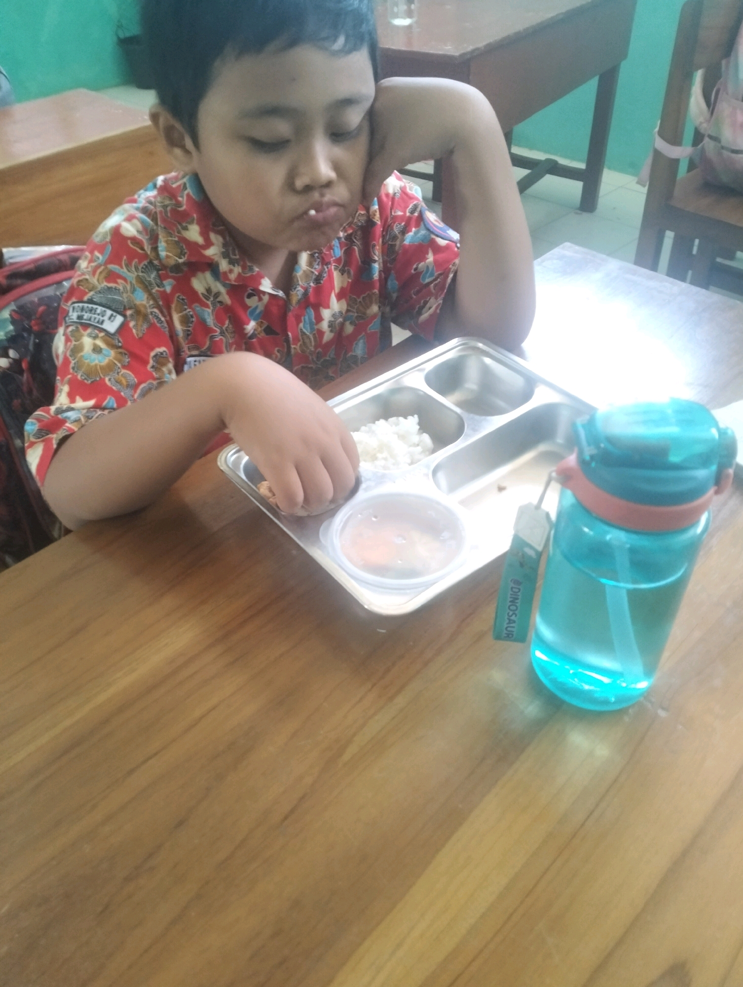 In Blabakan, Indonesia, a young boy is seated at a wooden desk, his arm resting on his cheek as he eats from a compartmentalized lunch tray. The tray contains white rice and what appears to be a small portion of fried food. A blue water bottle with a straw is placed next to the tray. The boy is wearing a red and white batik-patterned shirt. The background shows more wooden desks and chairs, suggesting a classroom setting.