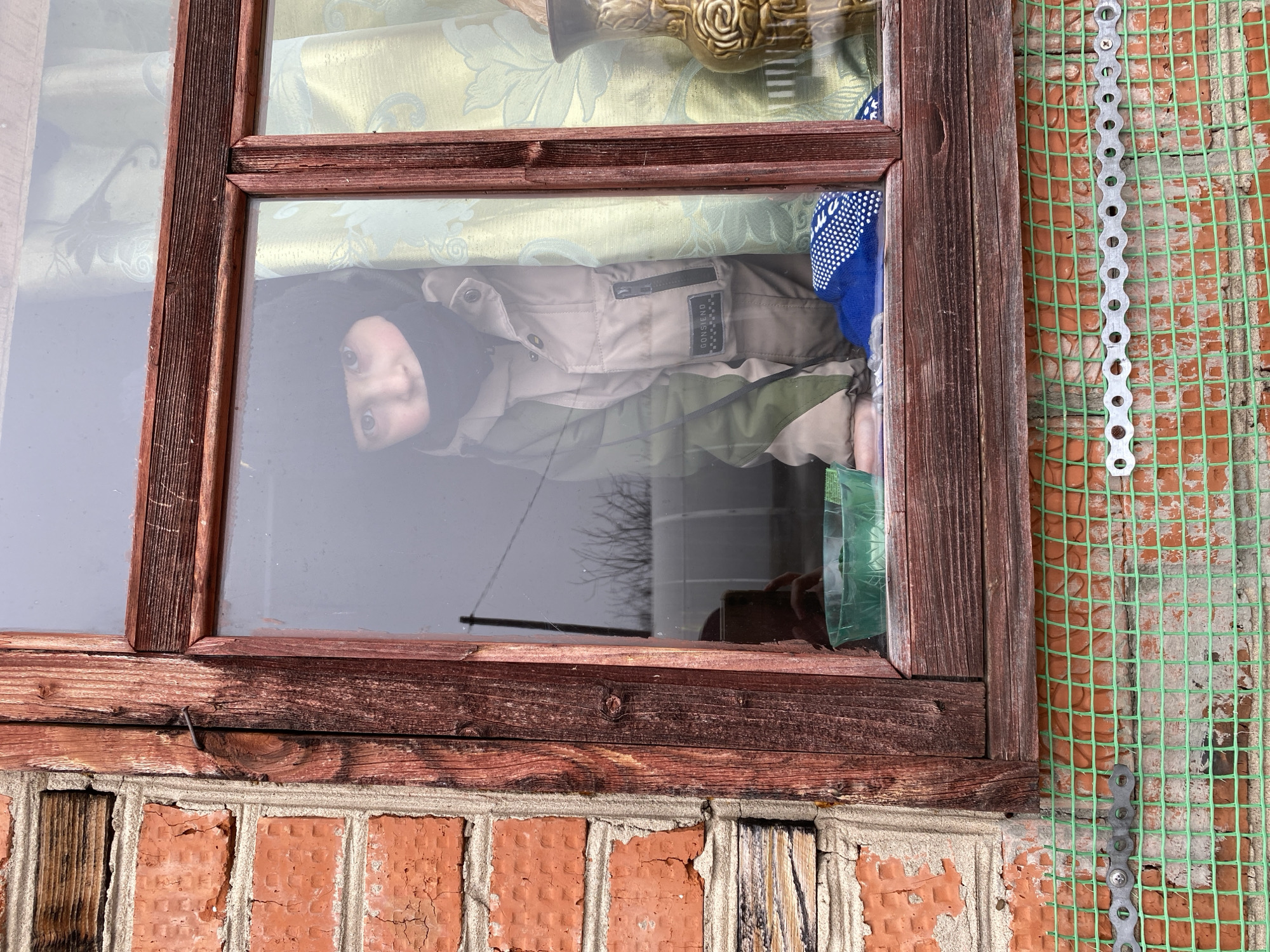 A child is looking out of a window in a residential building in what appears to be the city of Chaikovsky, Russia. The window frame is made of dark wood. The exterior wall of the building is constructed of red brick, with a green mesh overlay secured by a perforated metal strip. The child is wearing a dark head covering, a light tan jacket, and a blue patterned garment. The jacket has a label on it that reads "Godsieno". Inside, behind the child, is a patterned curtain and what appears to be a decorative vase. The scene suggests an ordinary moment captured within a domestic setting.