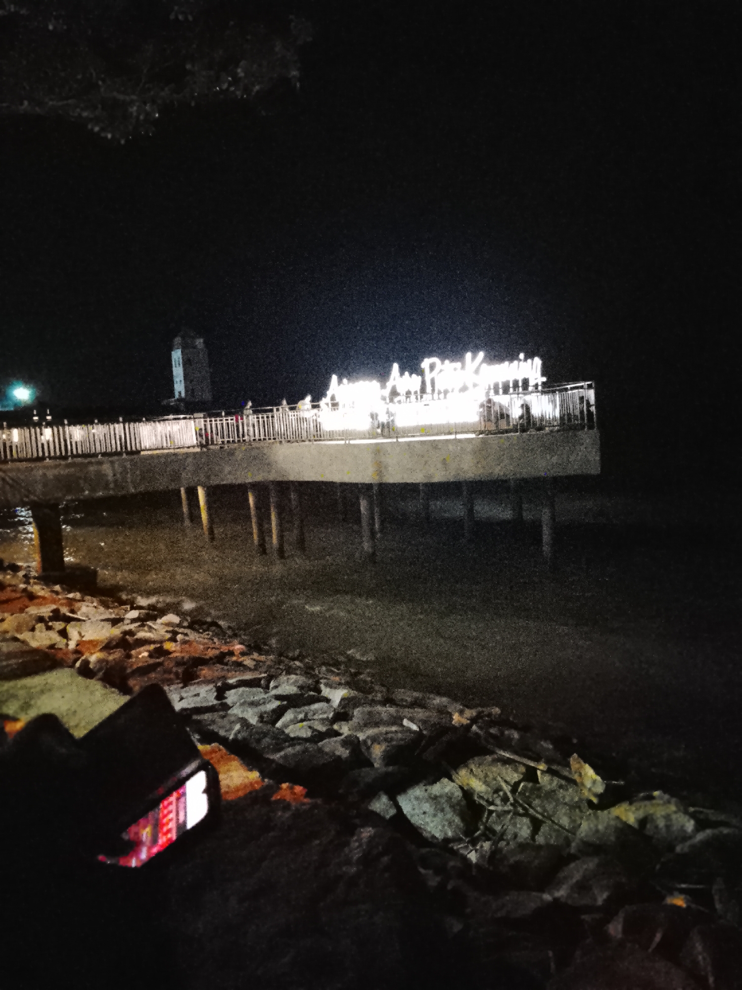 A night scene captures a pier in Tanjung Balai Karimun, Indonesia, illuminated by neon lights. A tall, light-colored tower with horizontal dark bands stands to the left of the pier. The pier itself has a light-colored railing with vertical white posts and is supported by thick, dark pilings that extend into the water. Several people are visible on the pier. A prominent neon sign on the pier reads "AYU PITA KEMUUN". In the foreground, a dark object, possibly a phone, rests on a bed of rocks near the water's edge, displaying a red and white illuminated screen. The water is dark and appears to be a shallow bay or estuary. The overall lighting is dim, with the pier and sign being the primary light sources.