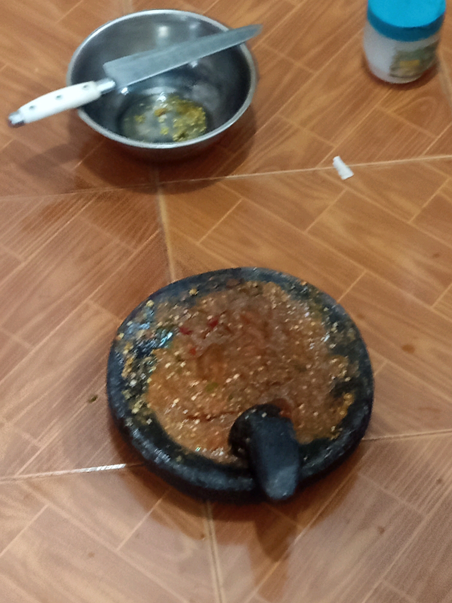 The image depicts a scene on a tiled floor, possibly in a kitchen or outdoor cooking area in Pulung Kencana, Indonesia. In the foreground, a traditional stone mortar and pestle is filled with a reddish-brown paste, likely a sambal or chili paste, with visible pieces of chili and other ingredients. The mortar has a worn appearance, suggesting frequent use.
In the mid-ground, a stainless steel bowl contains a small amount of a green, granular substance, possibly chopped herbs or spices, with a knife resting inside the bowl. The knife has a white handle. Further to the right, a small white plastic object, possibly a cap or tip, lies on the floor. A partially visible white jar with a blue lid is also present in the background.
The floor is covered with rectangular, wood-patterned tiles arranged in a herringbone pattern. The lighting suggests an indoor setting with overhead light, casting subtle shadows. There are no people visible in the image.