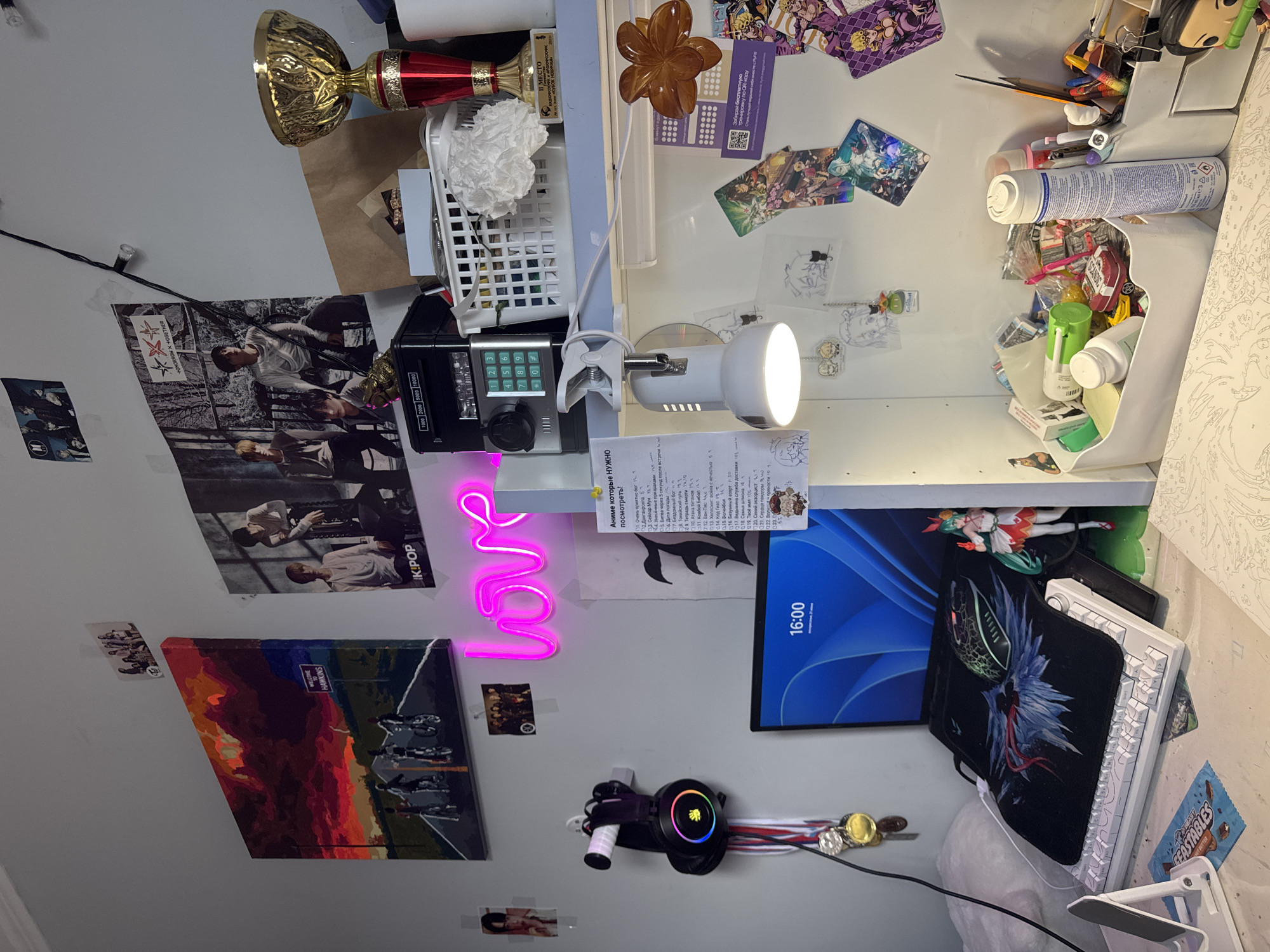 The image shows a desk area in a room, likely a teenager's bedroom. On the wall, there are several K-pop posters and a painting of a road with silhouettes of people on bicycles, under a dramatic sky. A pink neon sign that says "love" is also visible on the wall. A pair of headphones with colorful lights hangs on a hook, along with several medals.
On the desk, a laptop is open, displaying the time "16:00". A keyboard and mouse are set up for use, with a mousepad featuring a dragon design. A figurine of a character with green hair and a red and white dress stands to the right of the laptop.
To the right of the desk, there is a shelf with various items, including a white basket filled with toiletries and other small objects, a gold trophy, and a black piggy bank with a keypad. A desk lamp is clamped to the edge of the shelf, pointing downwards. The wall is decorated with more K-pop photos and some anime-themed cards and keychains.
A box filled with art supplies, including pens, pencils, and a can of spray paint, is also on the shelf. A partially completed paint-by-numbers artwork is visible in the foreground of the desk. The overall impression is one of a personal workspace decorated with interests and hobbies.
The city of Zassovskaya, Russia, is a rural settlement in Krasnodar Krai, known for its agricultural activities. The provided image does not contain any specific elements that would definitively place it in Zassovskaya or suggest any connection to the region.