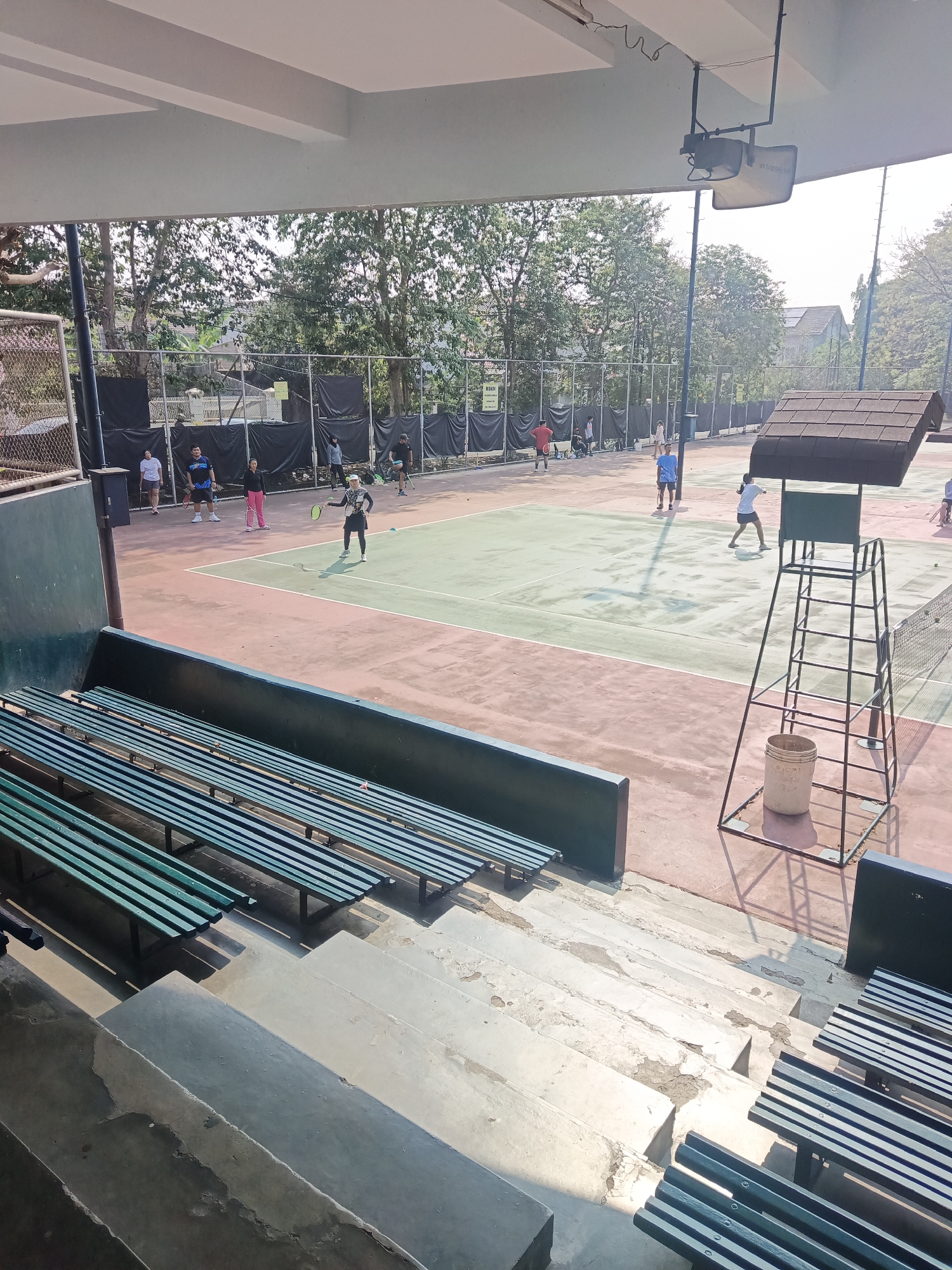 The image depicts an outdoor tennis court with several people playing tennis. The court appears to be made of a reddish-brown and green surface, with white lines marking the boundaries. A high umpire's chair with a brown roof stands to the right of the court. In the foreground, there are rows of green bleachers and concrete steps. Several trees and fencing surround the court, suggesting it is part of a sports complex. The lighting indicates it is daytime. The image was taken from an elevated position, possibly from a seating area or a building overlooking the court. Based on the presence of tennis courts and sports facilities, this location is likely a sports club or park within the city of East Jakarta, Indonesia.