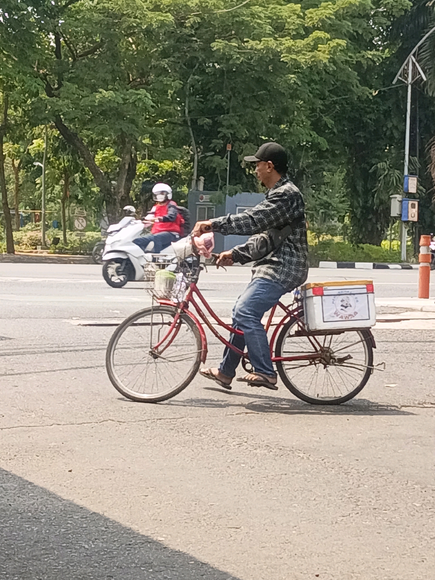 A man wearing a plaid shirt, blue jeans, and a black baseball cap rides a red bicycle with a white cooler box attached to the rear rack. He is on a paved road with trees and other vehicles in the background, including a white scooter with a rider in a red jacket and white helmet. The scene appears to be in an urban environment, possibly Surabaya, Indonesia, given the presence of tropical foliage and the style of vehicles.