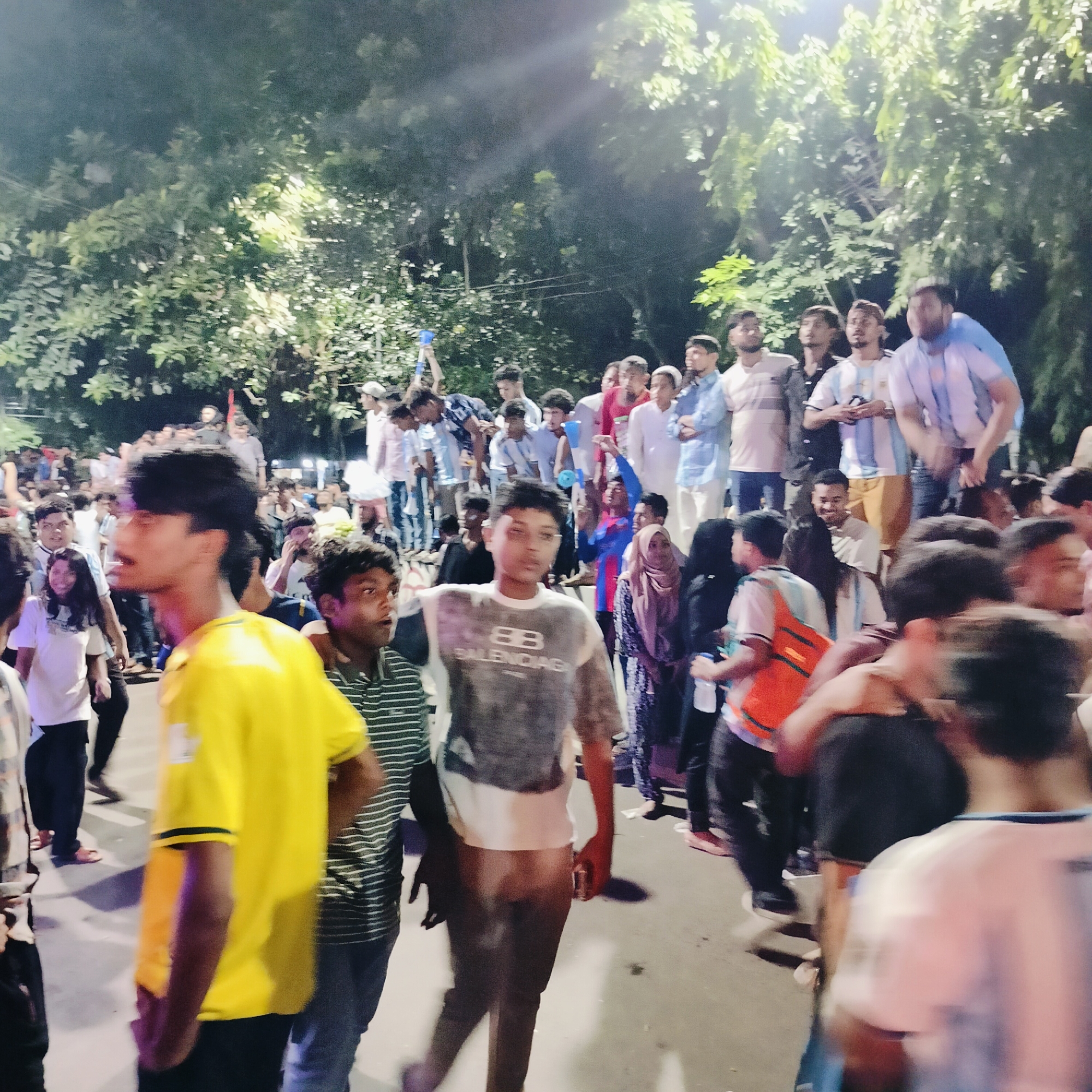 The image depicts a crowded street scene at night in Dhaka, Bangladesh, possibly during a sporting event or celebration, as many individuals are wearing what appear to be sports jerseys, including some in the colors of Argentina. The lighting is artificial, likely from streetlights and possibly some temporary lighting setups, casting a warm glow on the scene and creating shadows.
In the foreground, a young man wearing a bright yellow t-shirt with black trim is partially visible, facing to his left. To his right, another young man in a striped t-shirt is also visible. Behind them, the crowd is dense with people of various ages, mostly young adults, many of whom are looking in the same direction, suggesting they are watching something or participating in a collective activity. Some individuals are holding up flags or banners, and one person is holding a blue vuvuzela, a common instrument at sporting events.
The background features trees, indicating an outdoor urban environment. The atmosphere appears lively and energetic, with a sense of shared excitement among the people. The ground is paved, suggesting a street or public walkway. The image is slightly blurry, which is common in low-light, dynamic crowd shots, and adds to the sense of movement and excitement. The overall impression is one of a public gathering in a city setting, likely in a popular area of Dhaka.